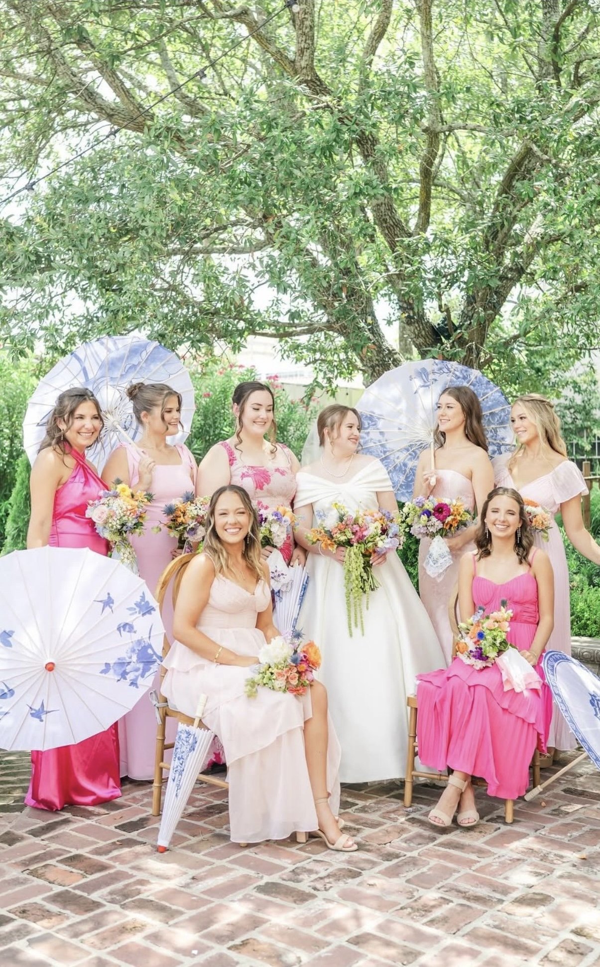 A group of women in colorful dresses holding flowers and parasols, posing outdoors under a large tree.