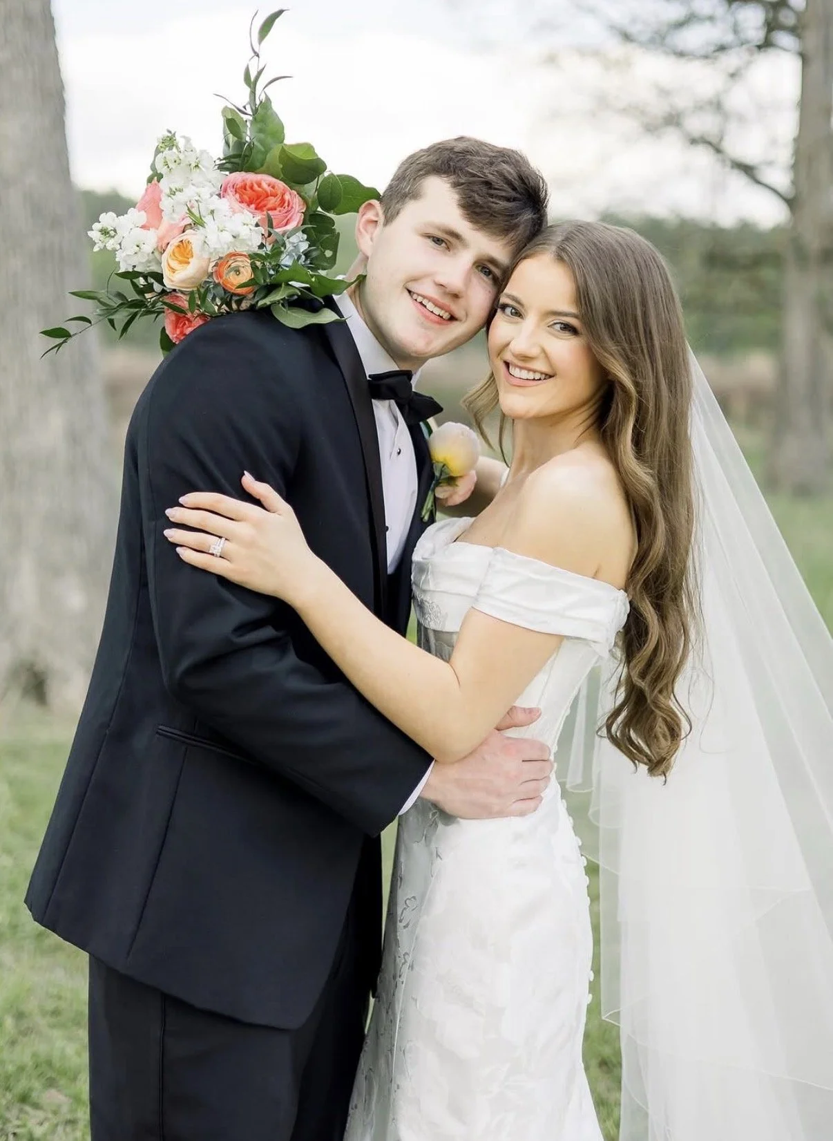 A bride and groom are embracing outdoors, smiling at the camera. The groom is wearing a black tuxedo with a bow tie and has a bouquet of flowers on his back. The bride is wearing an off-the-shoulder wedding dress and has a long veil.
