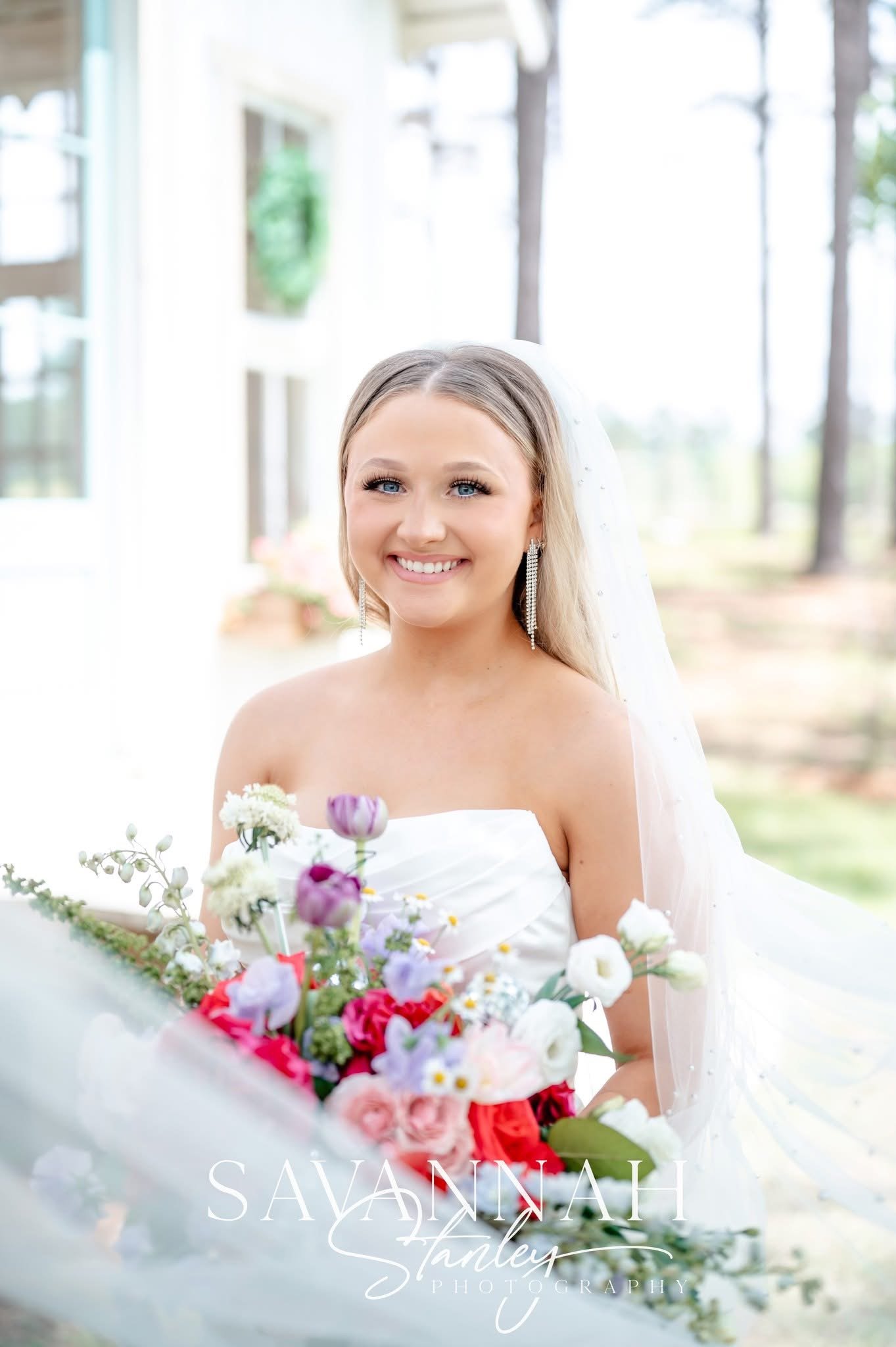Bridal portrait of a smiling blonde woman in a white wedding dress, holding a colorful bouquet of flowers, outdoors with a bright, natural background.