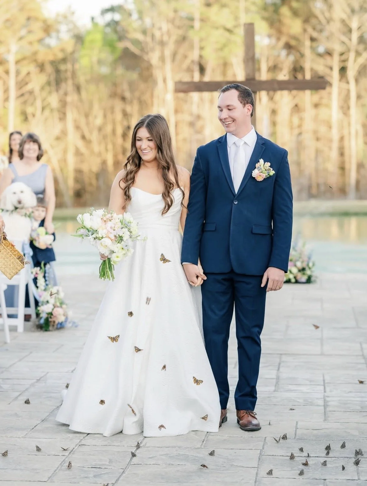 A bride and groom holding hands during an outdoor wedding ceremony. The bride is wearing a white wedding dress with butterfly decorations and holding a bouquet of flowers. The groom is dressed in a dark blue suit with a white shirt and tie. They are 