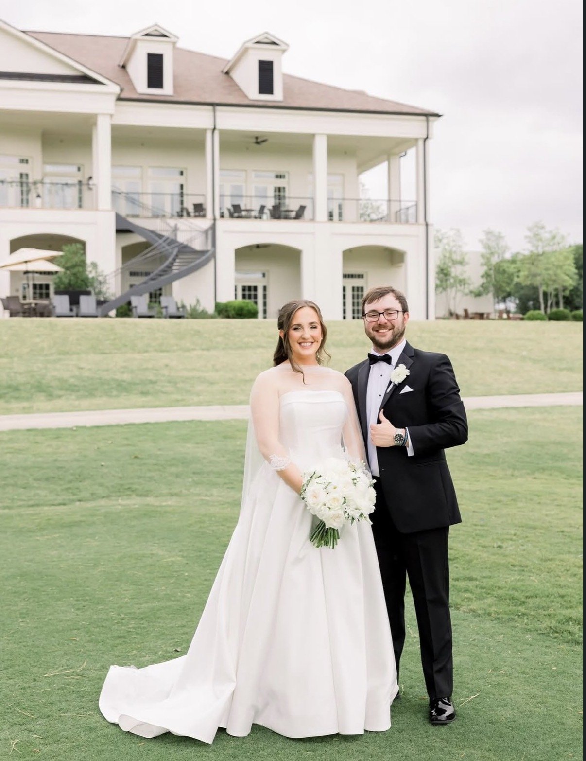 A bride and groom standing outdoors on a wedding day, smiling, with a large house in the background. The bride is holding a bouquet of white flowers, and the groom is dressed in a black tuxedo with a boutonniere.
