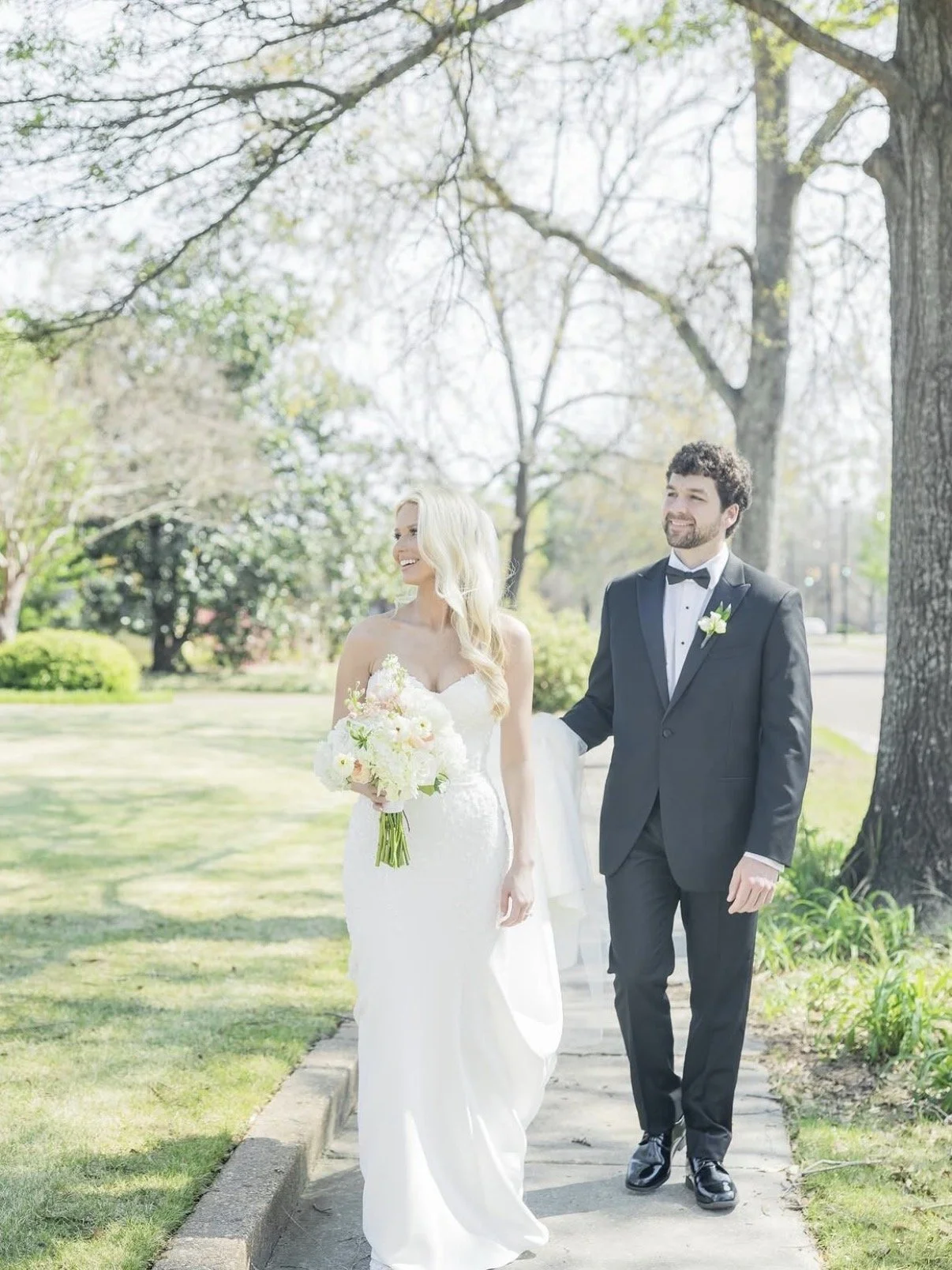 A bride and groom walking outside on a sunny day, smiling, with the bride holding a bouquet of white and pink flowers, and the groom in a black tuxedo with a bow tie and boutonniere, background of trees and greenery.