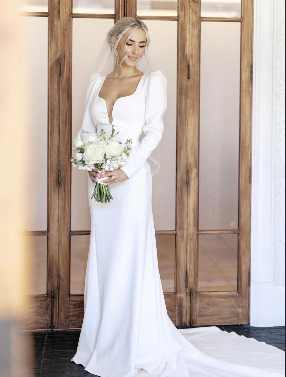 A bride in a white wedding dress holding a bouquet of white roses standing in front of a wooden partition.