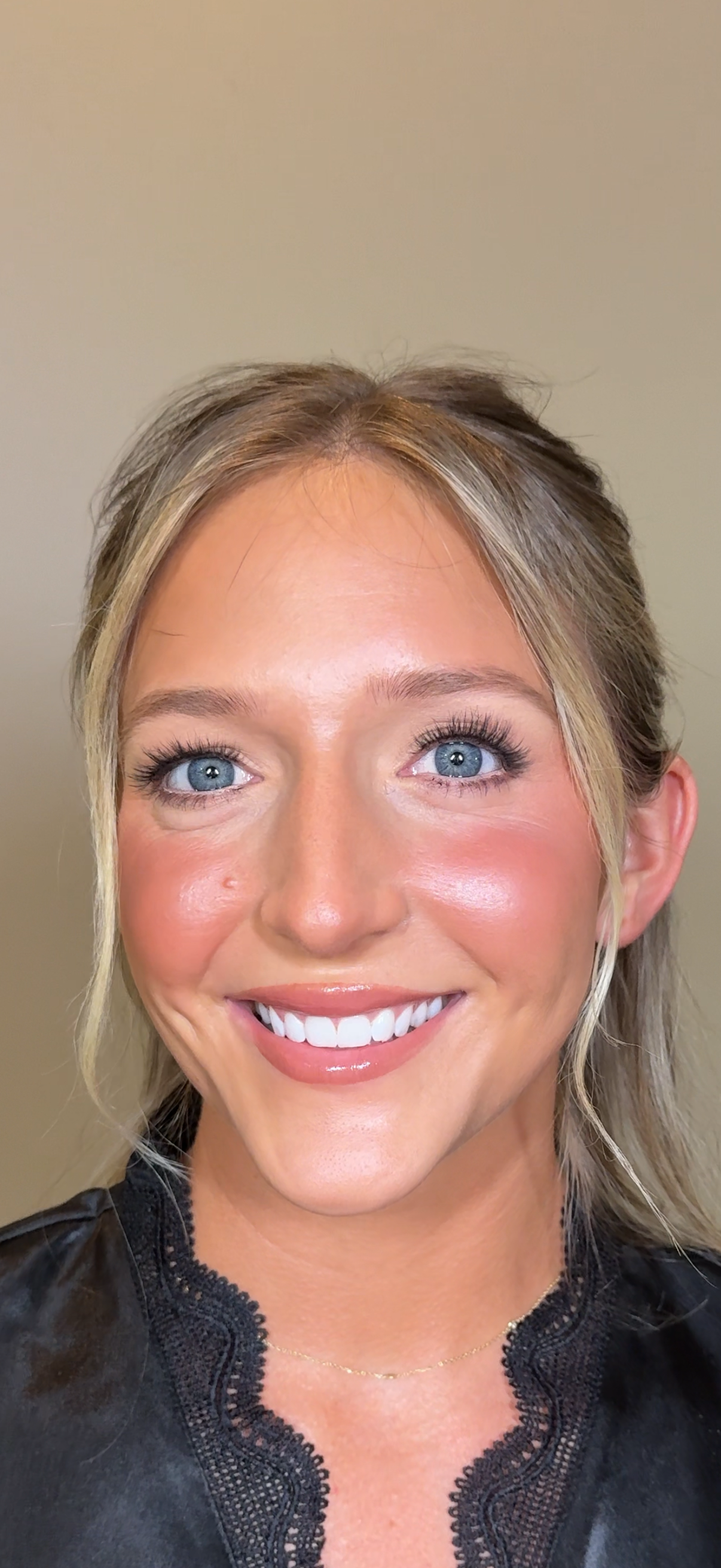 A smiling woman with blonde hair styled in loose waves, blue eyes, and natural makeup, wearing a black lacy top and a delicate gold necklace, standing against a plain beige wall.
