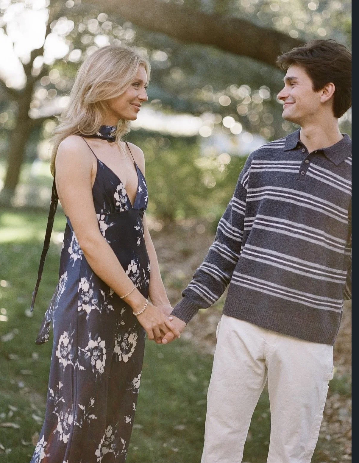 A woman and a man holding hands outdoors, smiling at each other. The woman is wearing a floral dress and the man is in a striped sweater and light pants. They are standing in a park with trees in the background.
