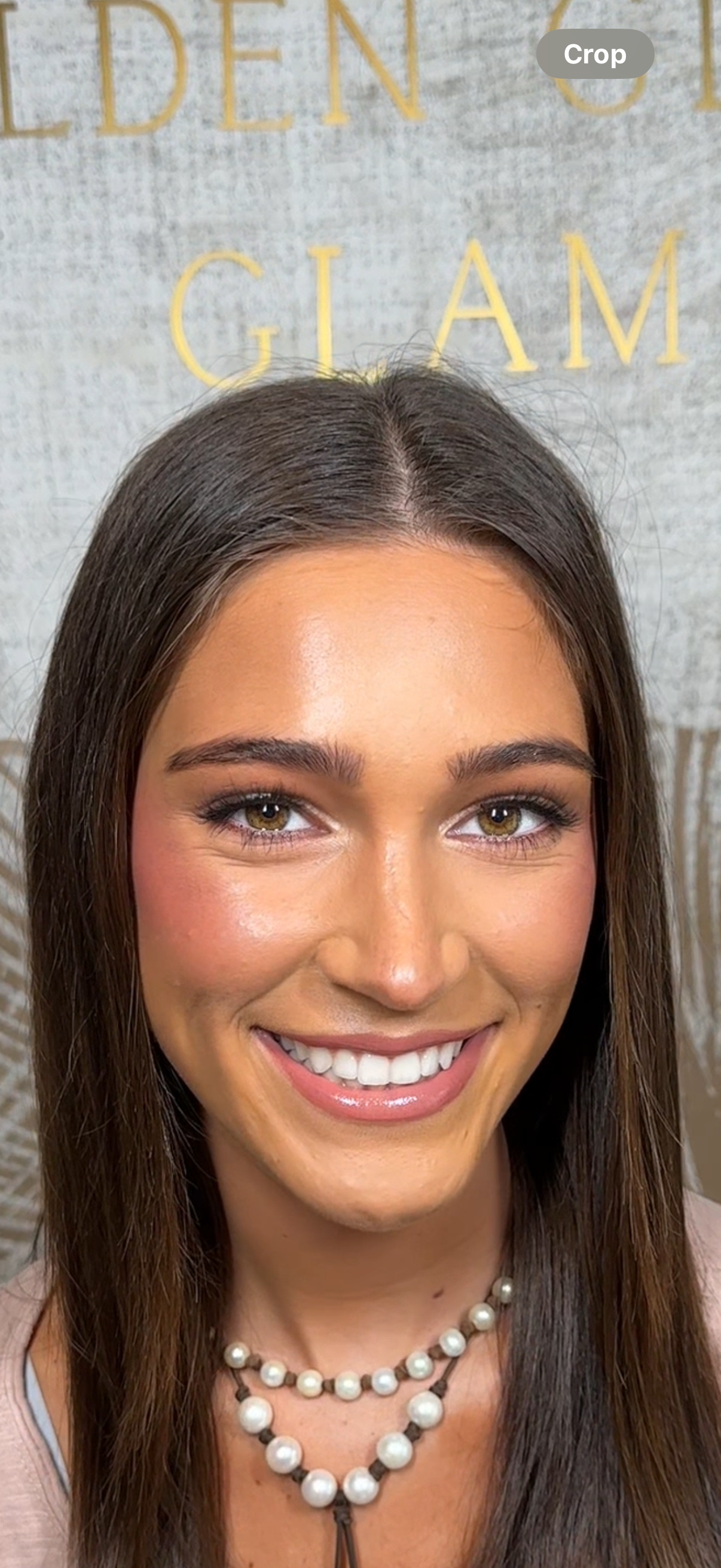 A woman with long brown hair and light makeup, smiling and wearing a pearl necklace, standing in front of a textured light gray wall with large yellow letters that say 'GLAM' and part of another word.
