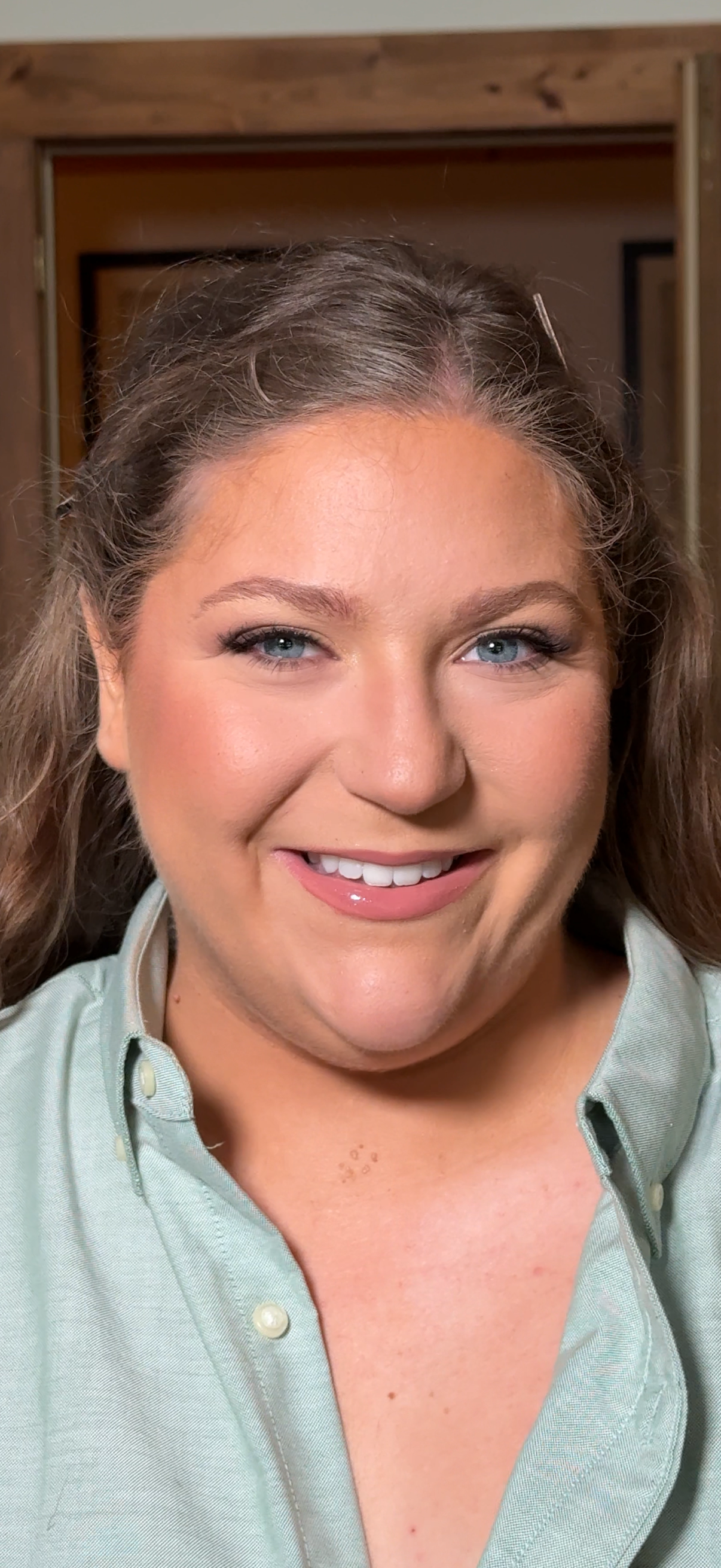 Close-up of a smiling woman with blue eyes and brown hair, wearing a light green button-up shirt, in an indoor setting with wooden elements in the background.