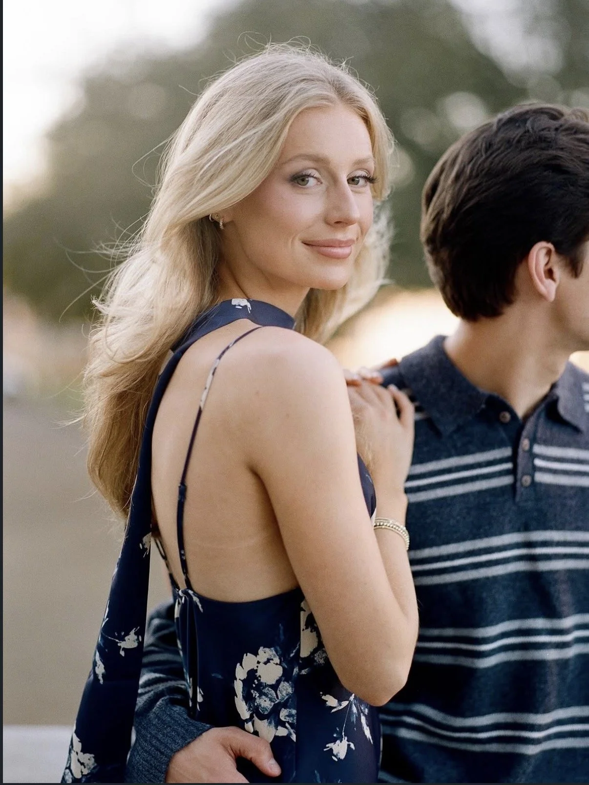 A woman with long blonde hair smiling while holding hands with a man in a striped shirt, outdoors with trees in the background.