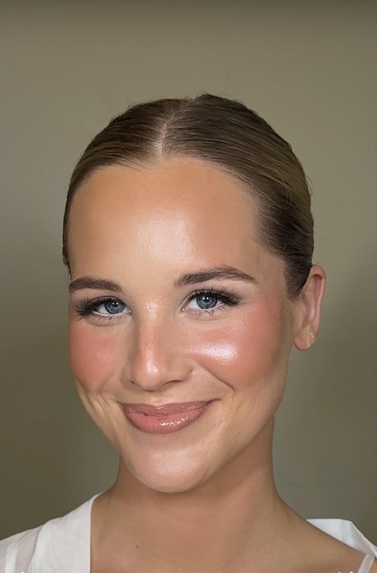 Portrait of a woman with fair skin, blue eyes, and brown hair styled in a sleek updo. She is smiling softly with makeup including eyeshadow, mascara, blush, and glossy lips. She is wearing a white top and a small earring.