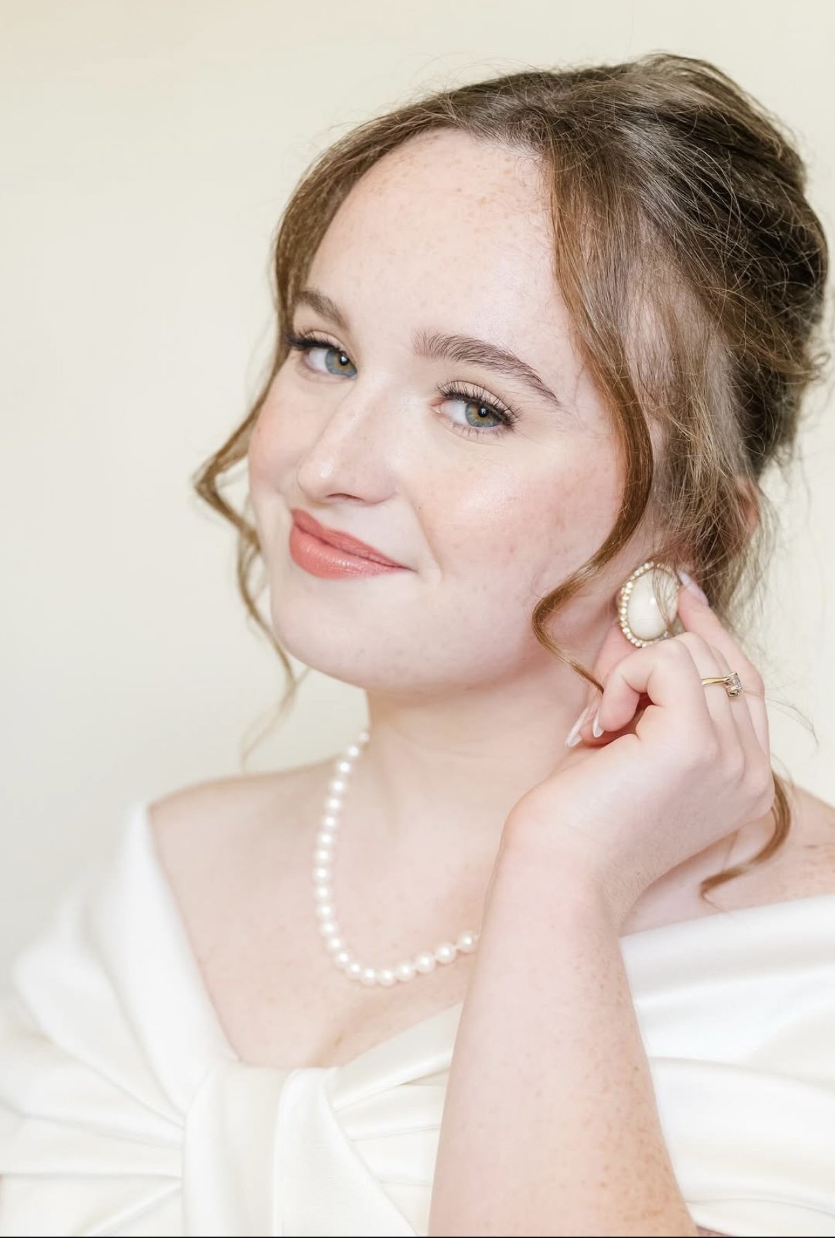 A woman with light skin and light brown hair styled in loose curls pinning back on one side, wearing pearl earrings, a pearl necklace, and a white dress, smiling softly and touching her ear.