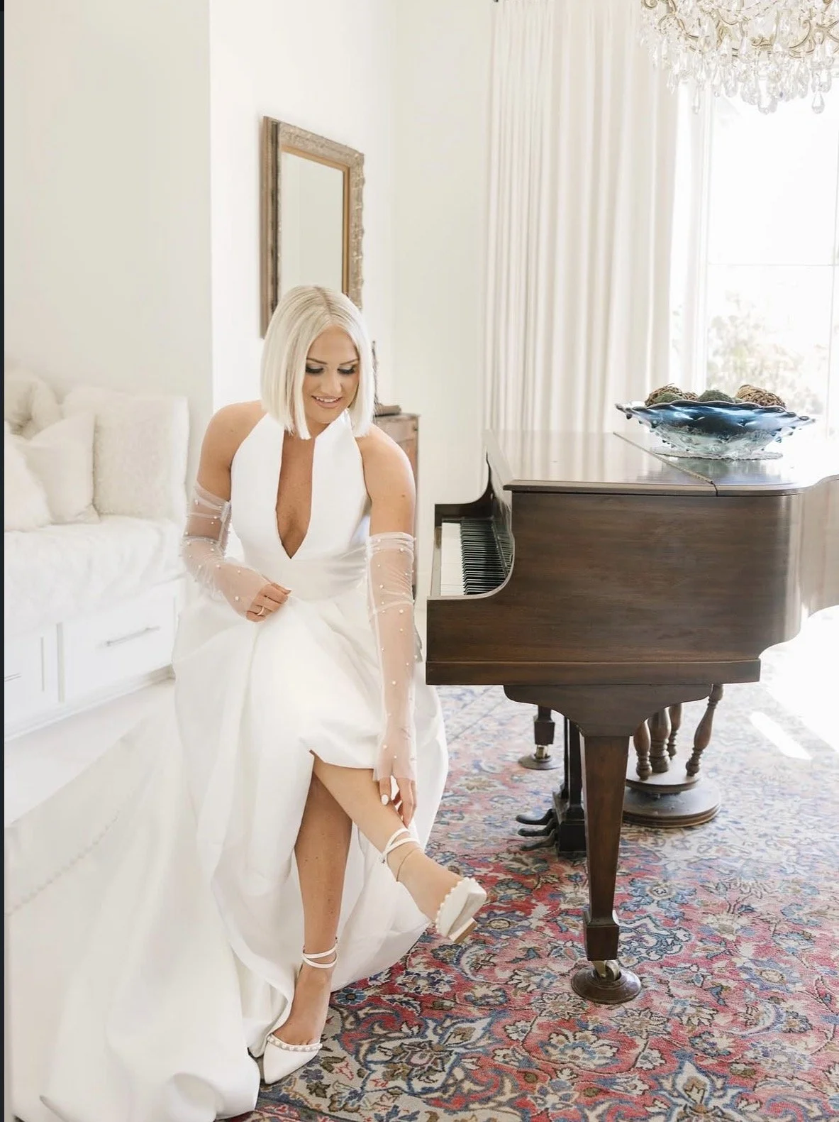A woman in a white dress sitting on a white sofa, lifting her right leg to show her shoe, next to a grand piano in a bright, elegant room with curtains and a chandelier.