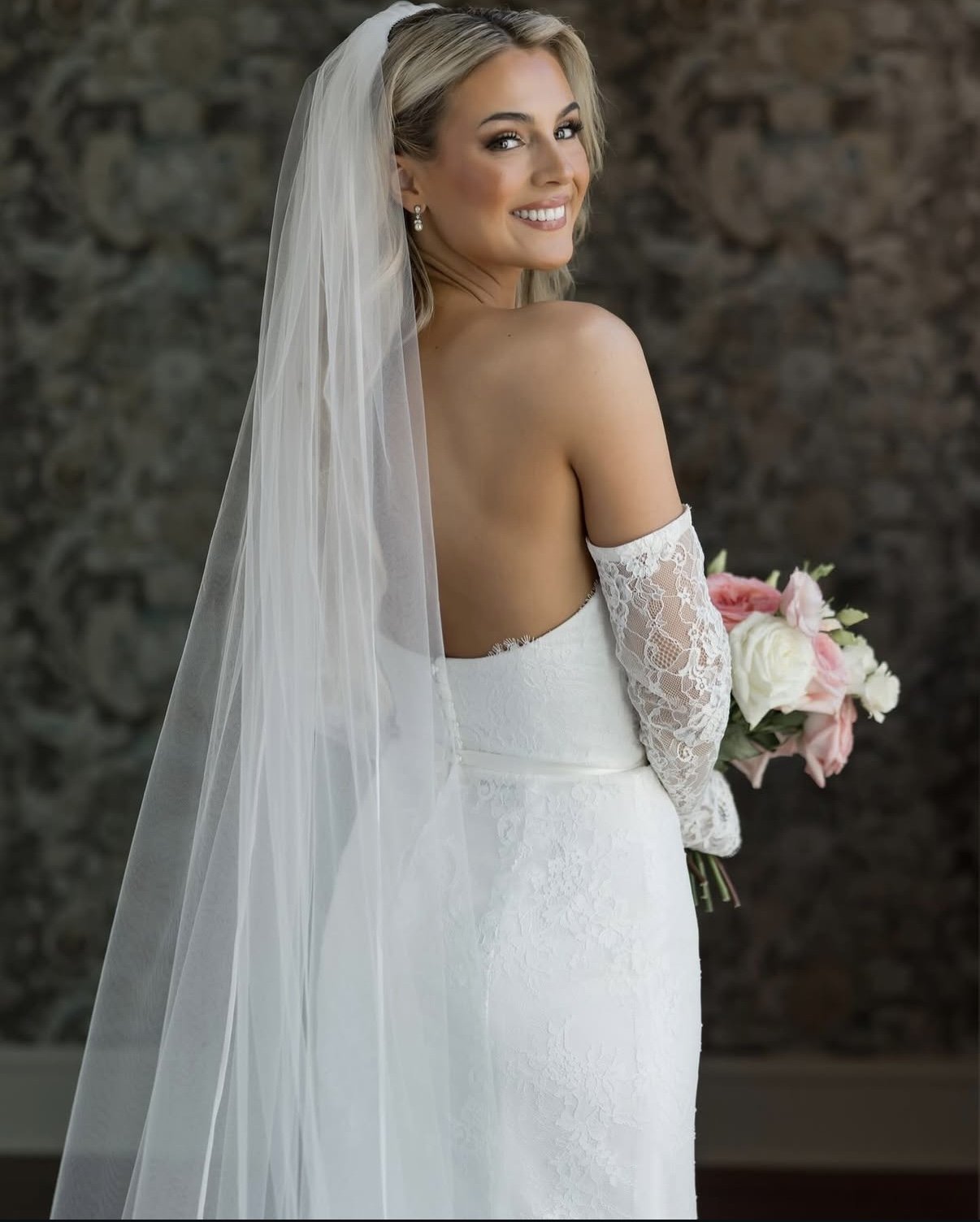 A smiling bride in a white wedding gown with lace off-the-shoulder sleeves and a long veil, holding a bouquet of pink and white roses, standing against a stone wall.