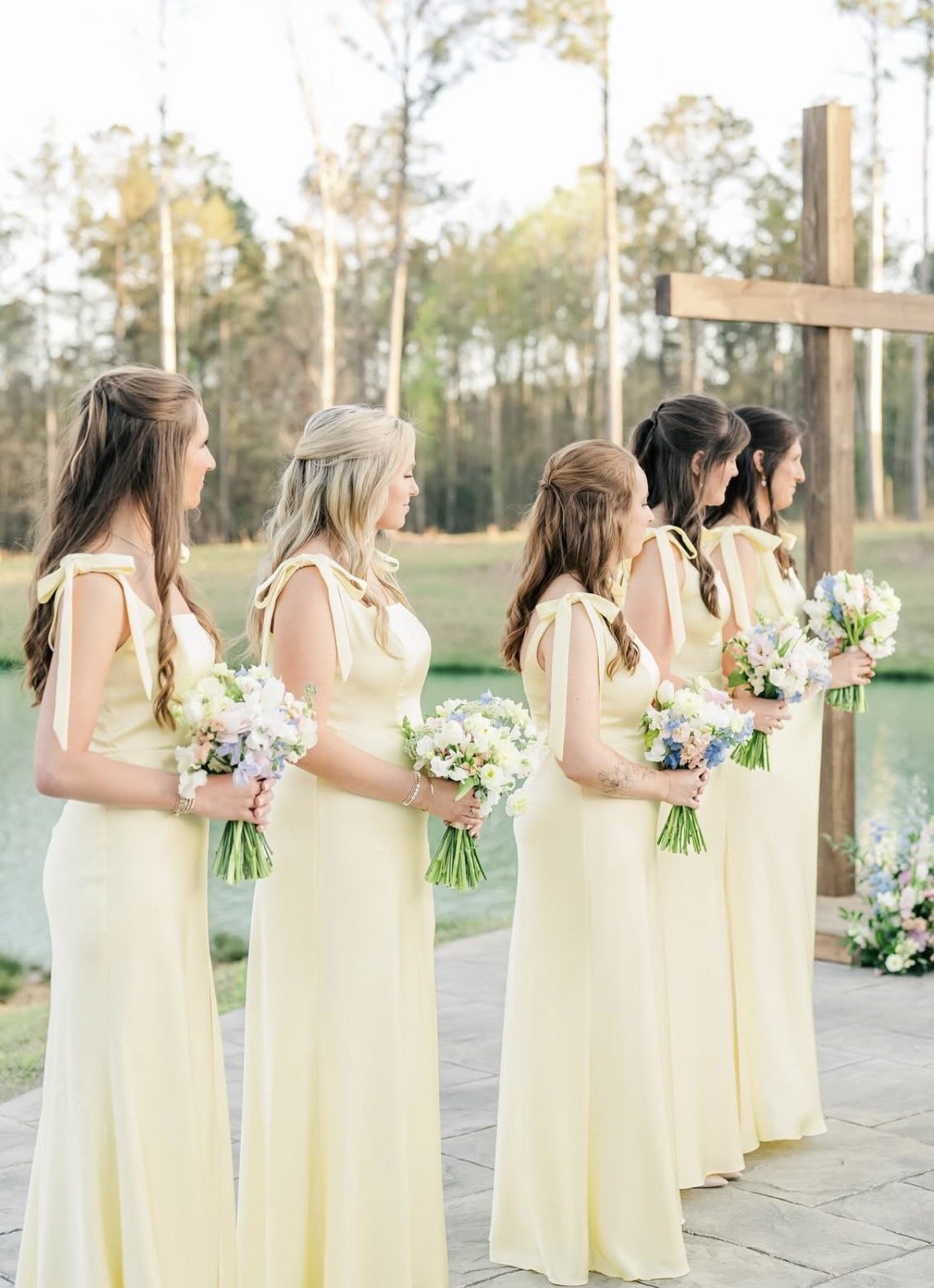 A group of five women dressed in long yellow dresses holding bouquets of flowers during a wedding ceremony outdoors near a lake and trees.