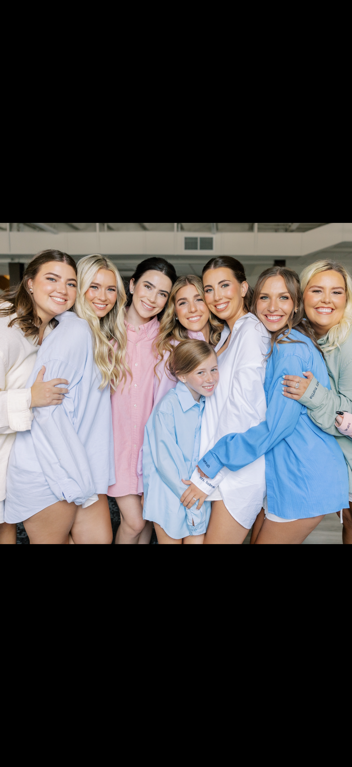 Group of smiling young women and a girl hugging each other, dressed in colorful oversized shirts, in an indoor setting.