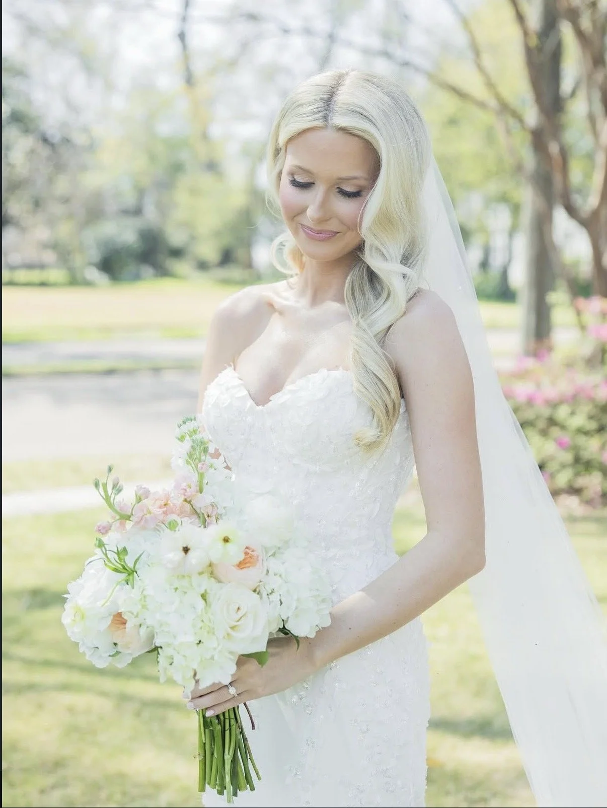 A bride with long blonde hair, wearing a white lace wedding dress with a sweetheart neckline, holding a bouquet of white and pastel flowers, outdoors in a park with trees and greenery in the background.