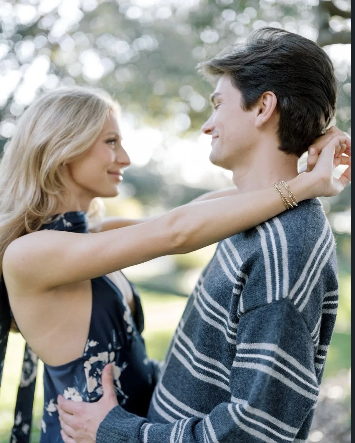 A couple embraces outdoors, smiling at each other, with the woman holding the man's neck and the man holding her waist, during daytime with blurred trees in the background.