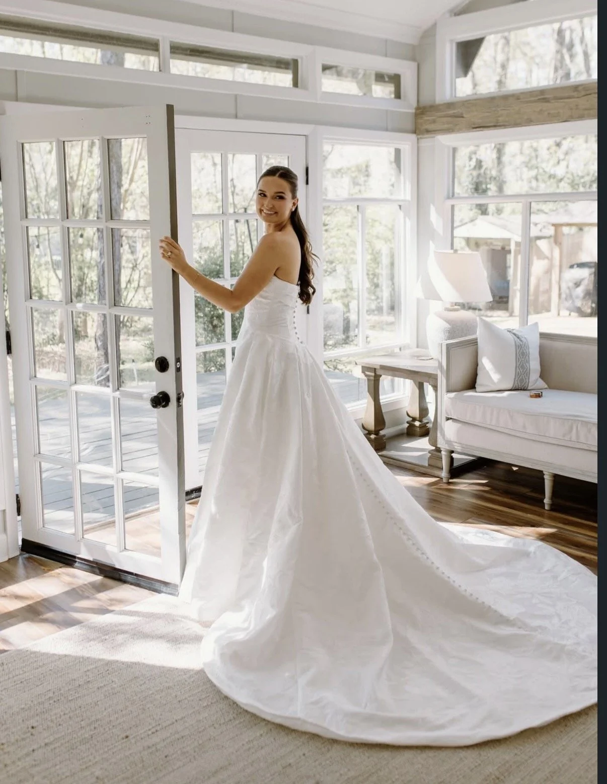 A woman in a white wedding dress standing in a bright room near a glass door, smiling, with large windows and a couch in the background.