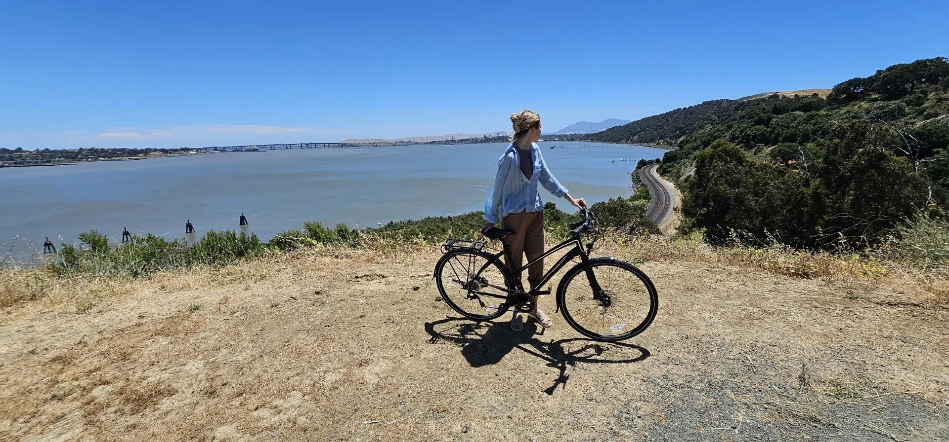 A woman standing with a bicycle on a dirt path overlooking a large river, with hills and a train track on the right and a bridge in the distance under a clear blue sky.