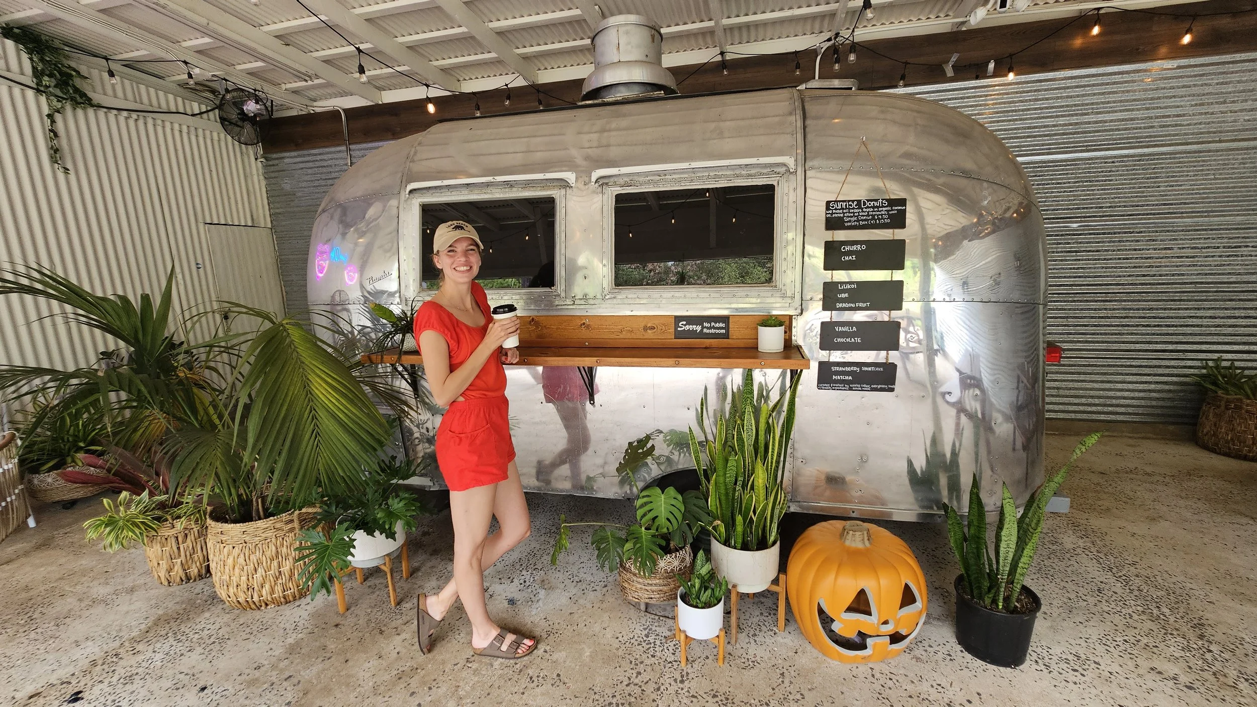 Smiling woman in red outfit holding coffee cup standing in front of vintage silver Airstream trailer serving as a food stand, decorated with potted plants, a pumpkin decoration, and menu signs.