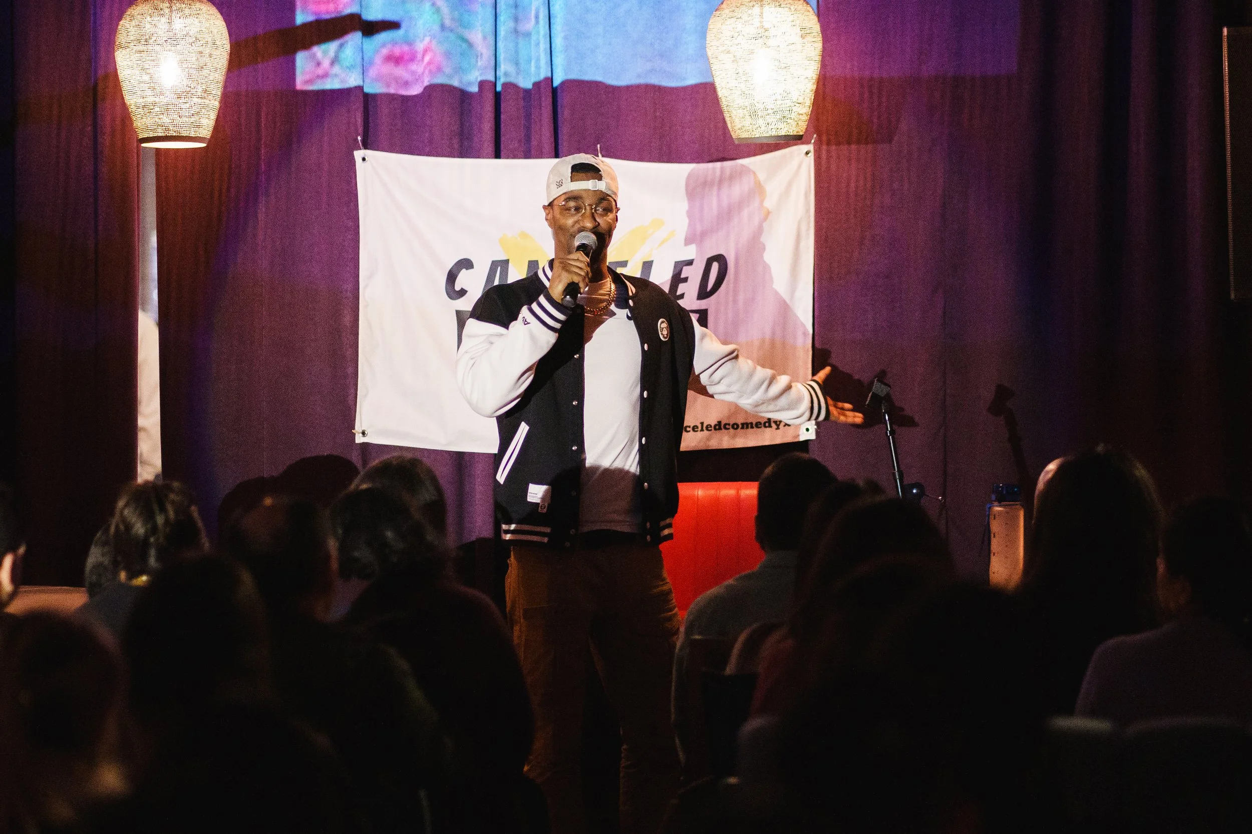 A man performs stand-up comedy on stage in front of a seated audience. He is wearing a varsity jacket, baseball cap, and glasses, holding a microphone. The backdrop has a white banner with the words "CANCELED" with some additional text, and two hanging light fixtures are above him.