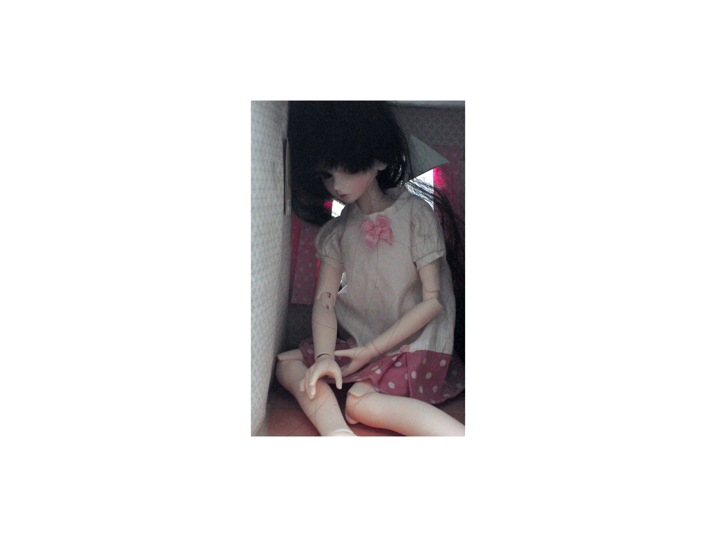 A large, melancholic doll with black hair sits crammed inside a small cardboard room, filling the entire space, creating a surreal and confined atmosphere.
