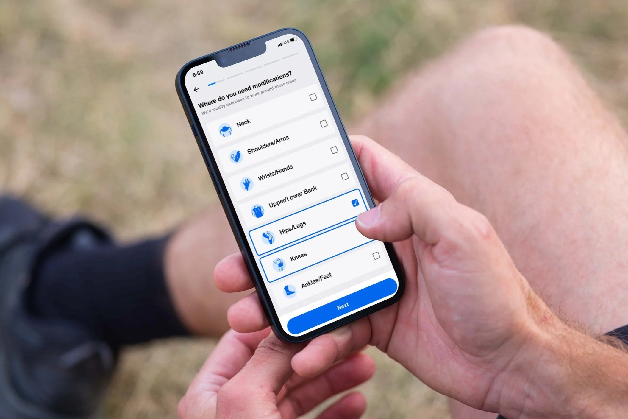 Person holding smartphone outdoors, viewing a health app for selecting body parts for exercise or injury modification, showing checkboxes for areas like hips, legs, knees, and ankles.