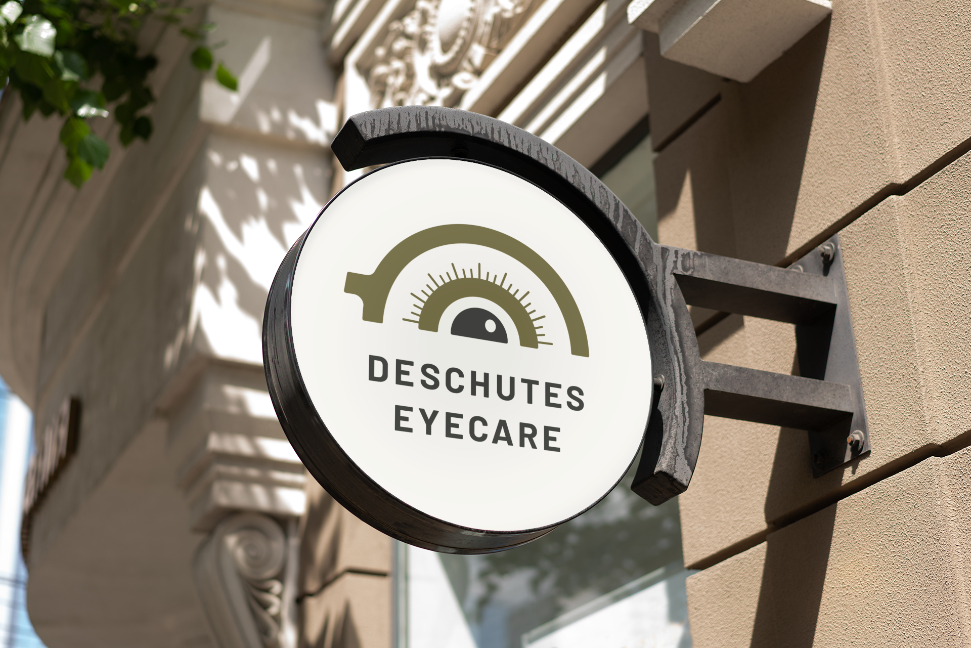 Outdoor sign for Deschutes Eyecare attached to a beige building wall, featuring a circular logo with an eye and gauge design.