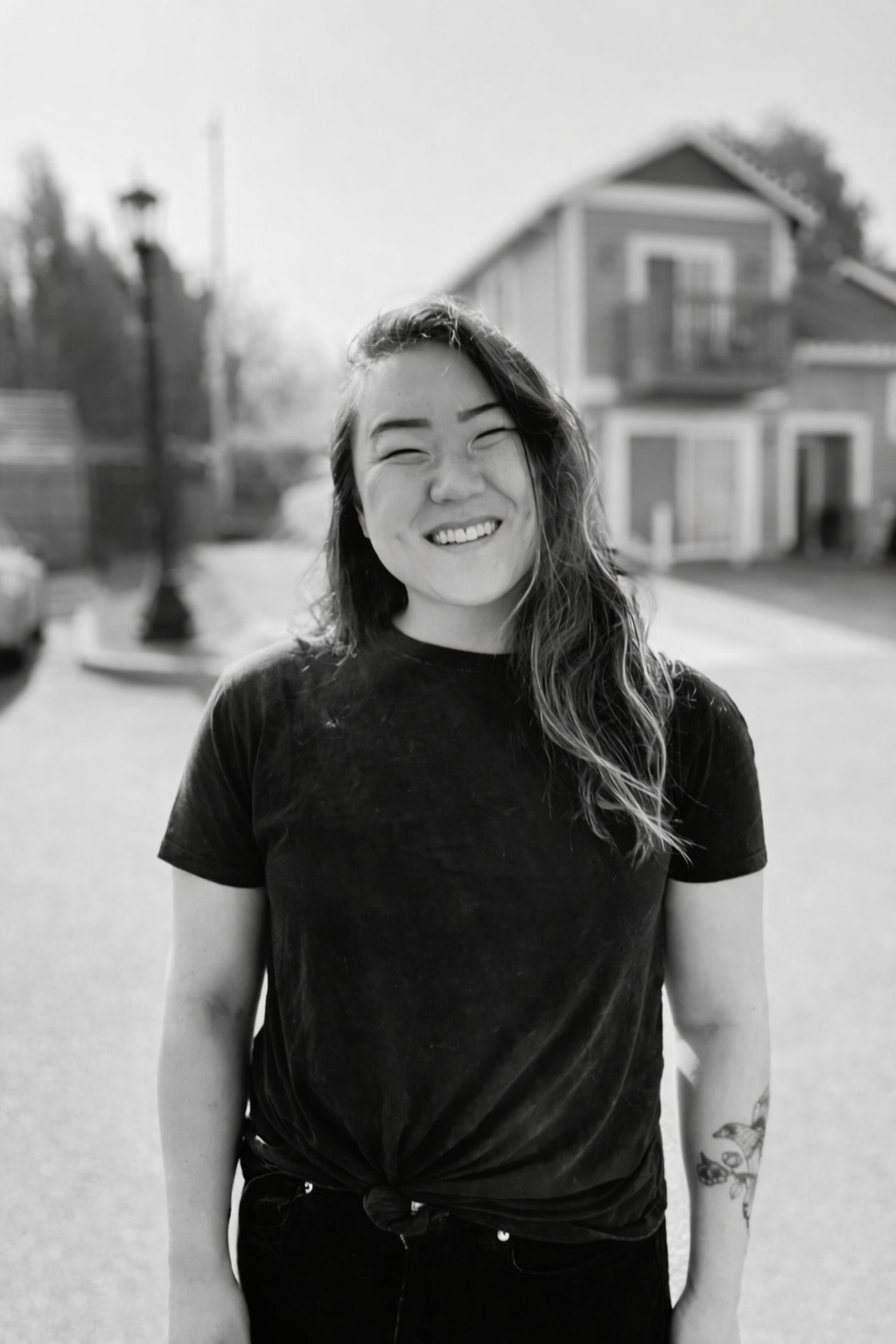 A young woman with long wavy hair, smiling and wearing a dark t-shirt, standing outdoors in a neighborhood with houses and trees in the background.