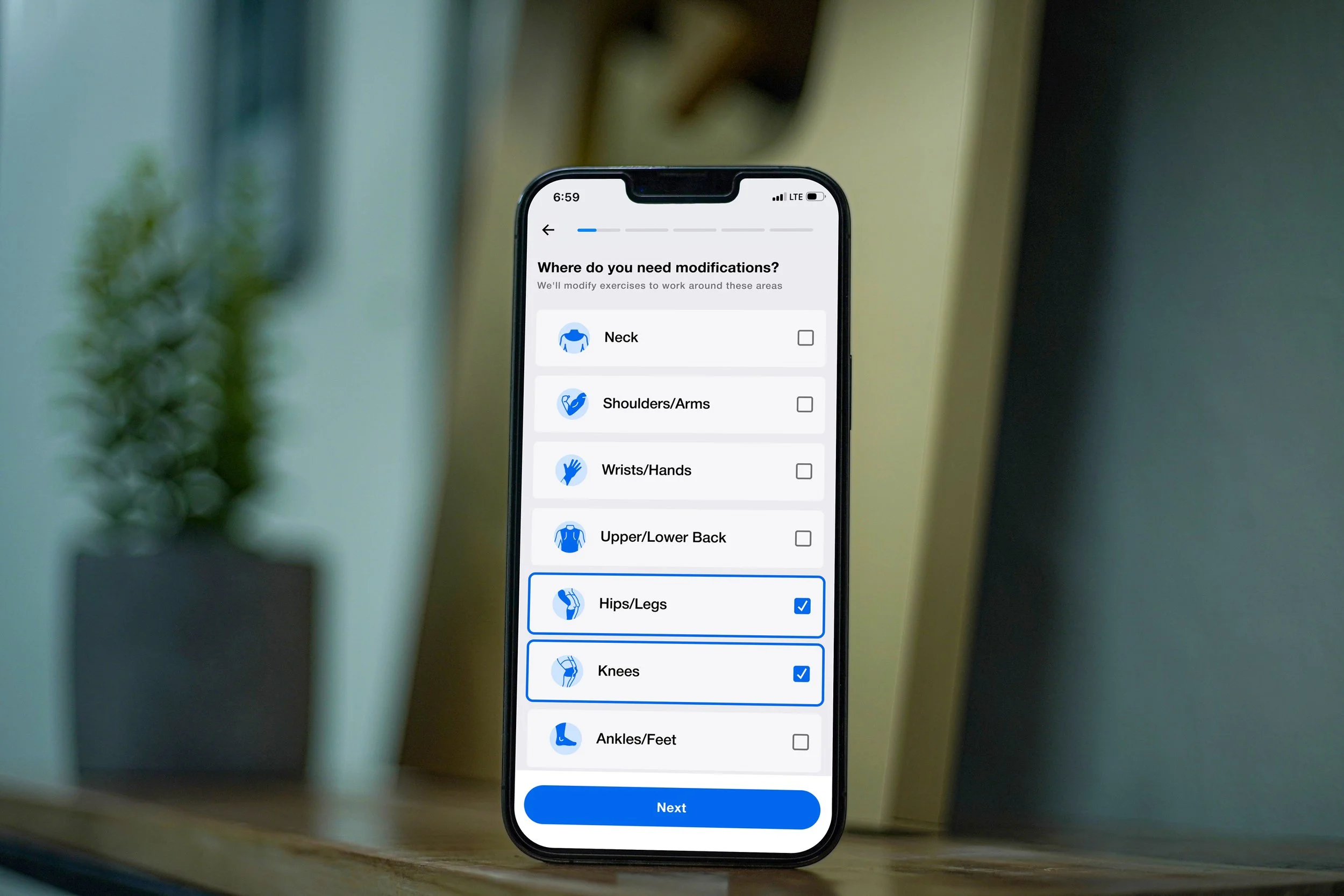 Smartphone displaying a fitness app with options to select areas for modifications such as hips and knees, with checkmarks indicating selections, placed on a wooden surface with a blurred potted plant in the background.