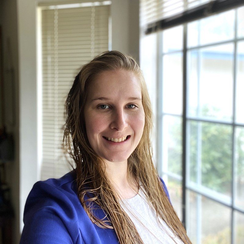 A woman with wet hair smiling indoors near a large window with white blinds, wearing a blue top.