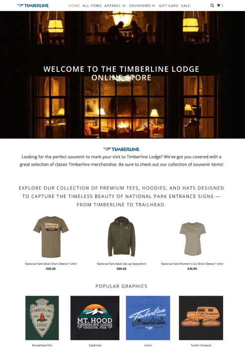 A cozy lit-up house at night with large windows showing people sitting inside, advertising Timberline Lodge merchandise, including T-shirts, hoodies, hats, and graphic designs related to national parks and mountain themes.