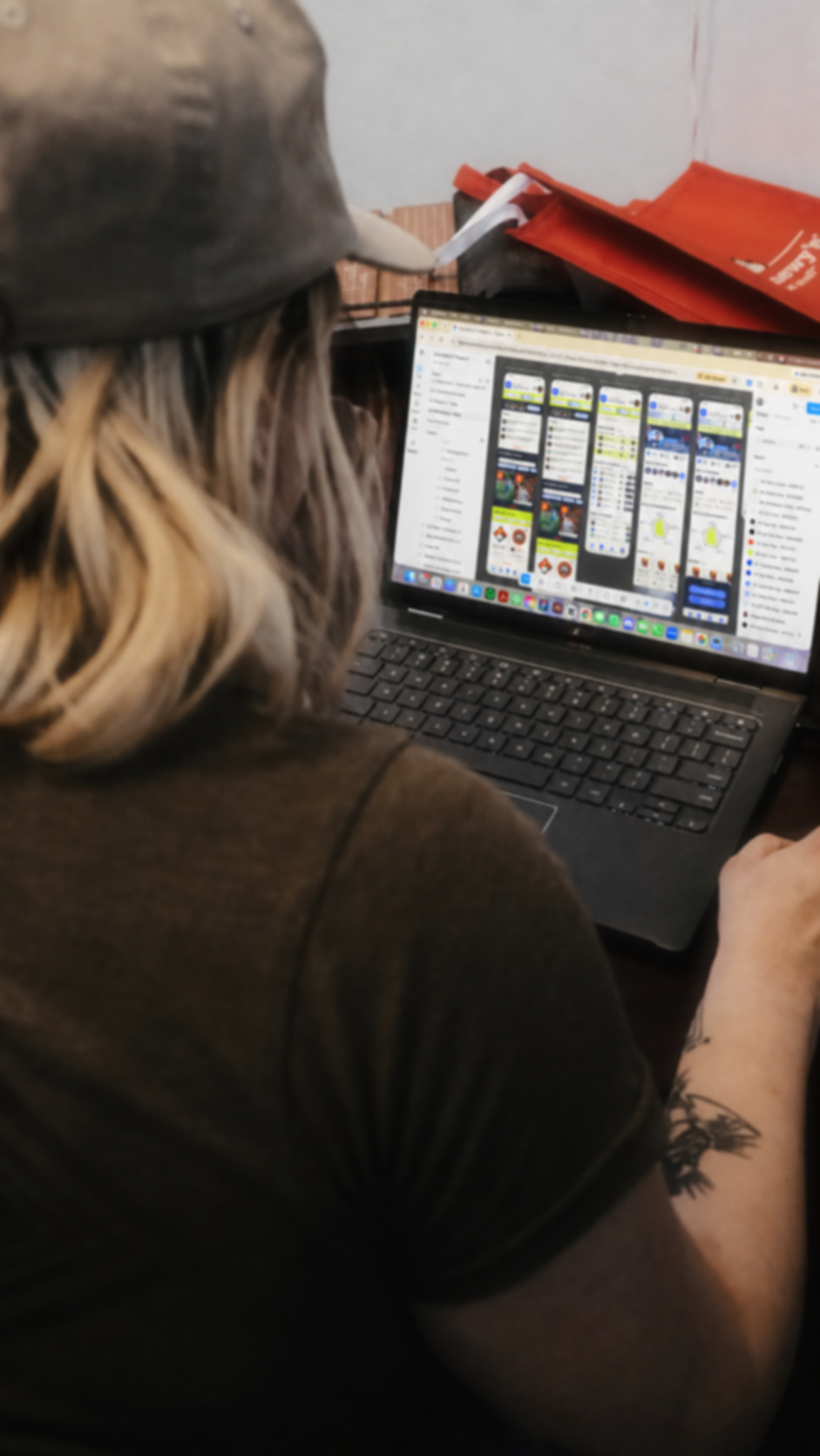 A person with long wavy blonde hair wearing a dark baseball cap and dark shirt using a laptop. The laptop screen displays a social media platform with multiple posts and images. The person has a tattoo on their right arm. There are orange bags and a white object on the surface behind them.
