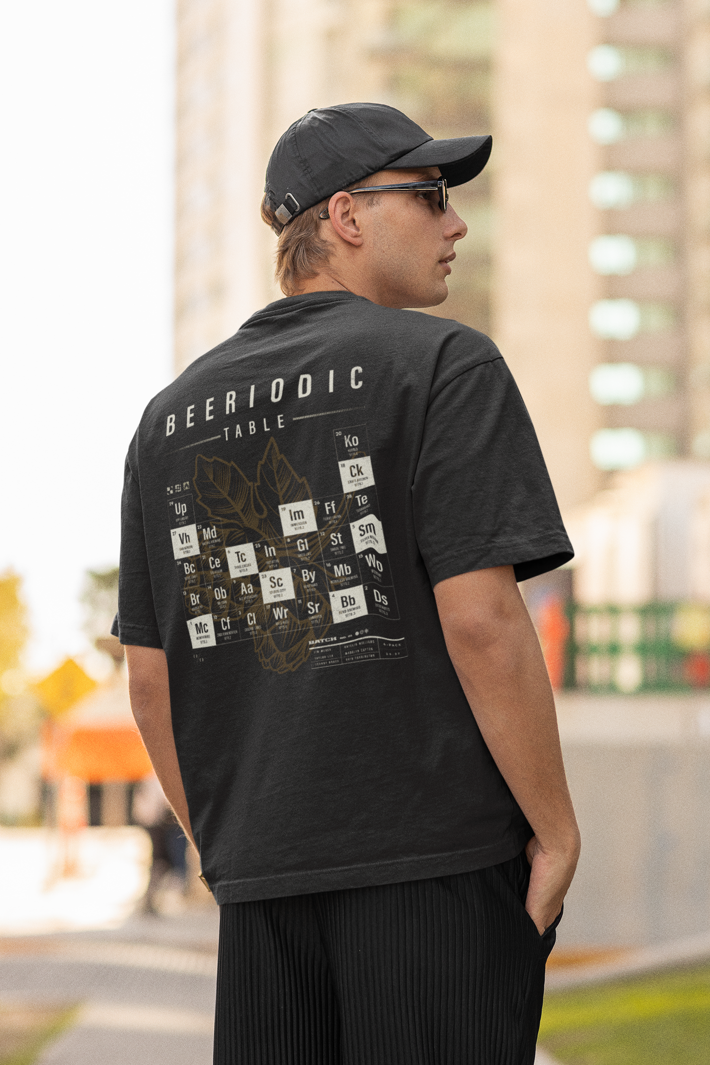 A young man standing outdoors in an urban setting. He is wearing a black cap, black sunglasses, a grayscale graphic t-shirt with the periodic table of elements and the word 'BEERIODIC,' and black pants. His right hand is in his pocket and he's lookin