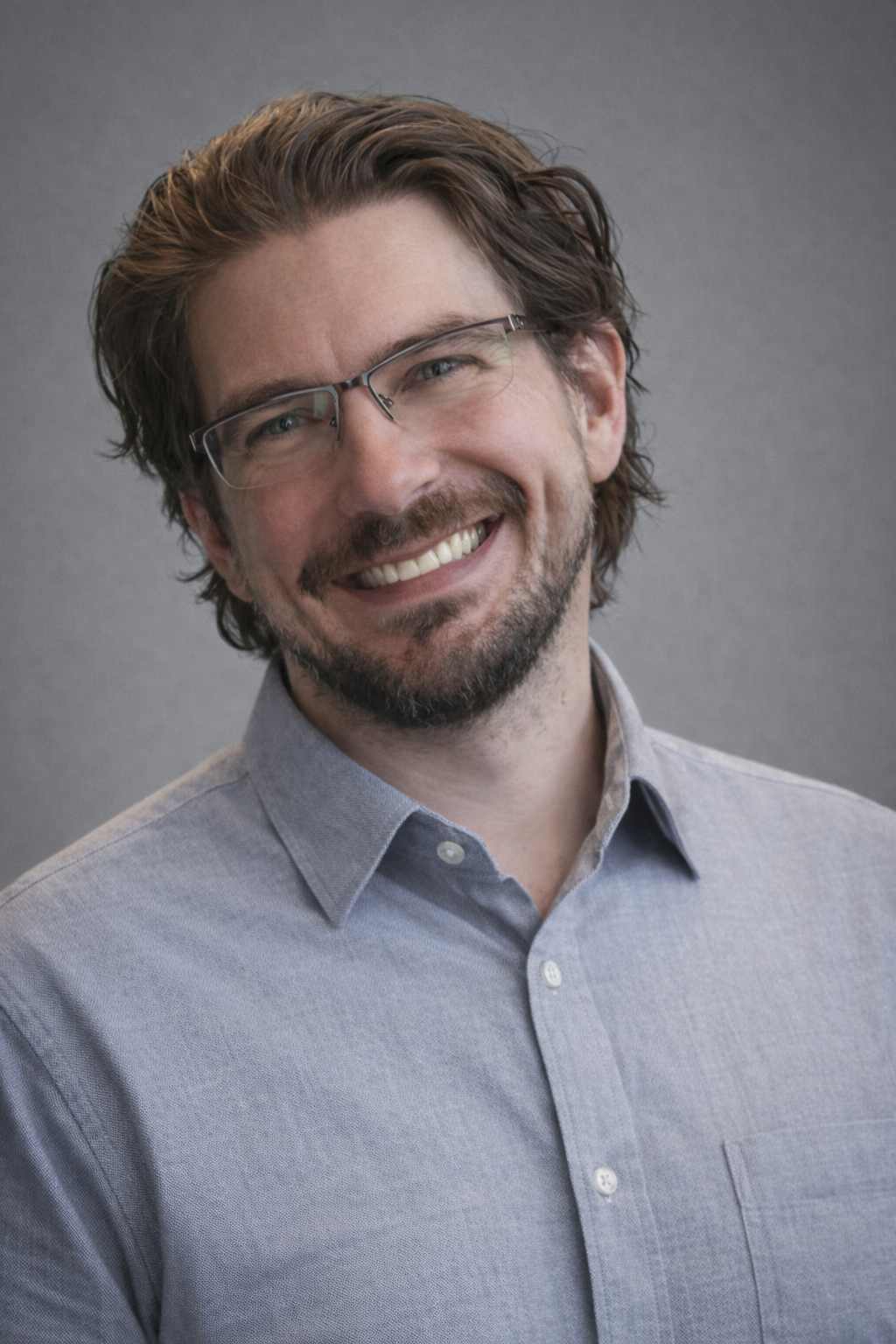 A smiling man with glasses, with brown hair and a beard, wearing a light gray button-up shirt, standing against a gray background.