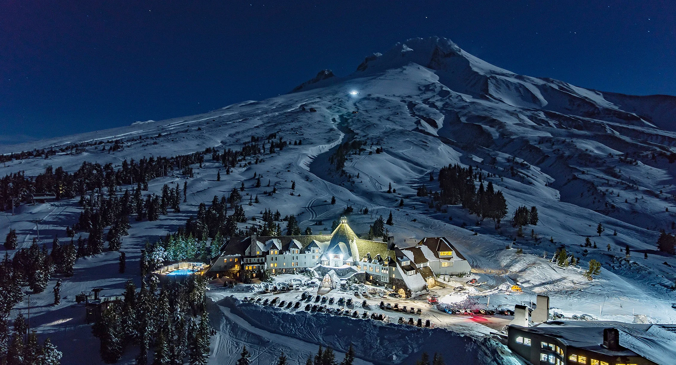 A snow-covered mountain at night with a ski trail illuminated by a bright light. At the base, there's a large, lit-up hotel or lodge surrounded by trees and parked cars.