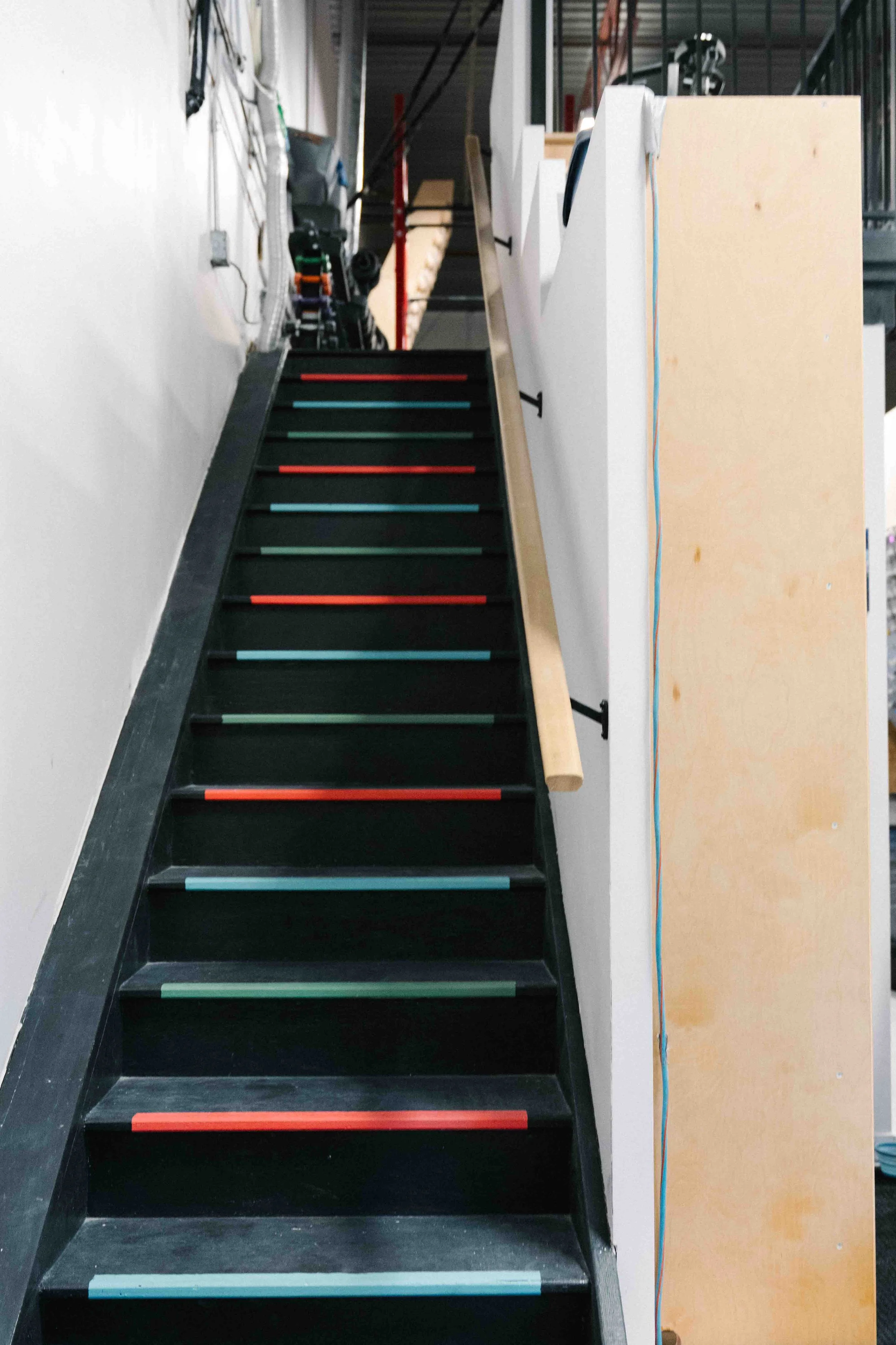 A staircase with black steps and colorful striped anti-slip strips on each tread, located inside a building with a white wall on the left and a beige wall with a blue wire on the right. The staircase leads upward to a higher level with visible metal 