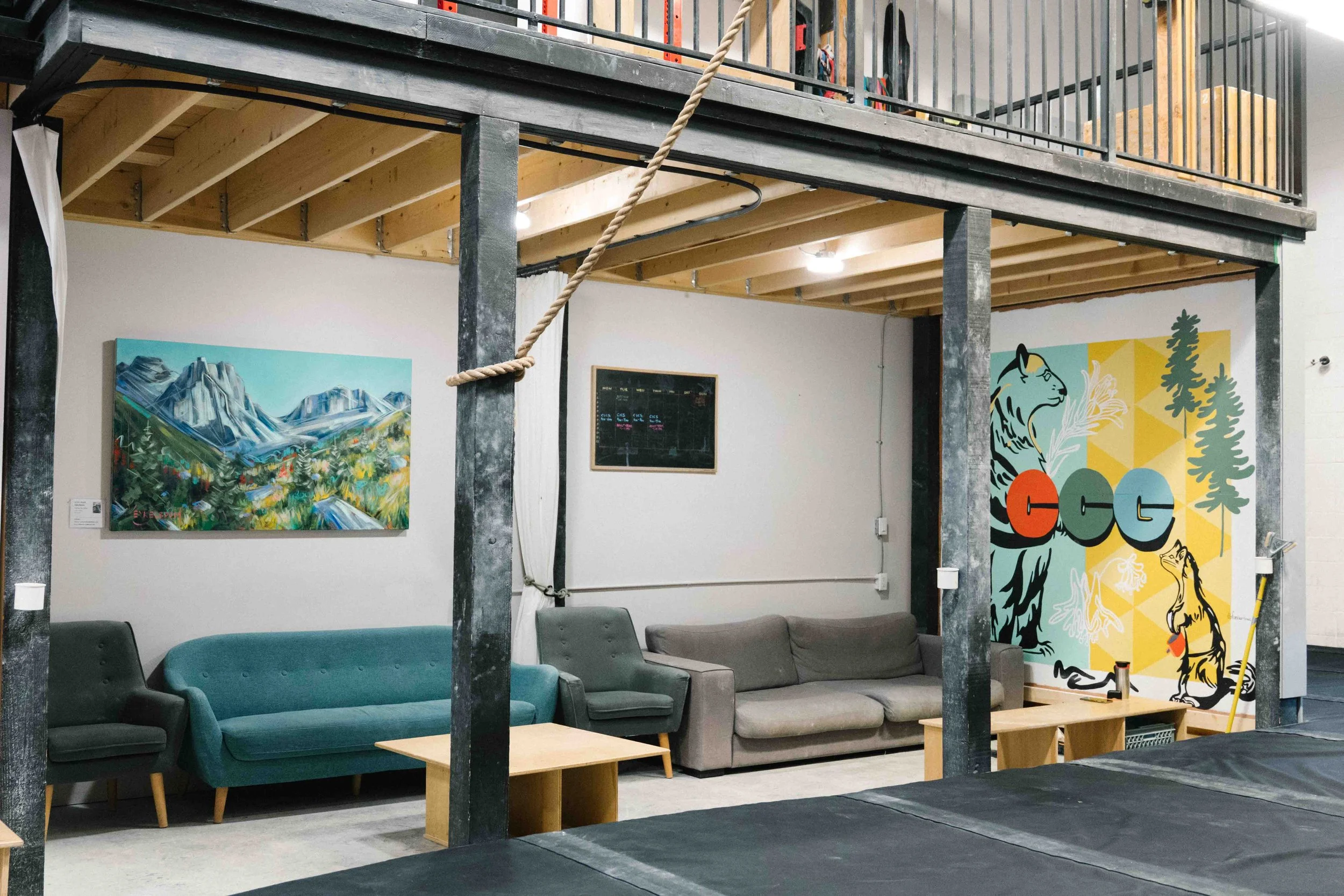 An indoor space with a lounge area, featuring a teal sofa and a gray sofa, wooden coffee tables, artwork of mountains and animals on the walls, and a black chalkboard.