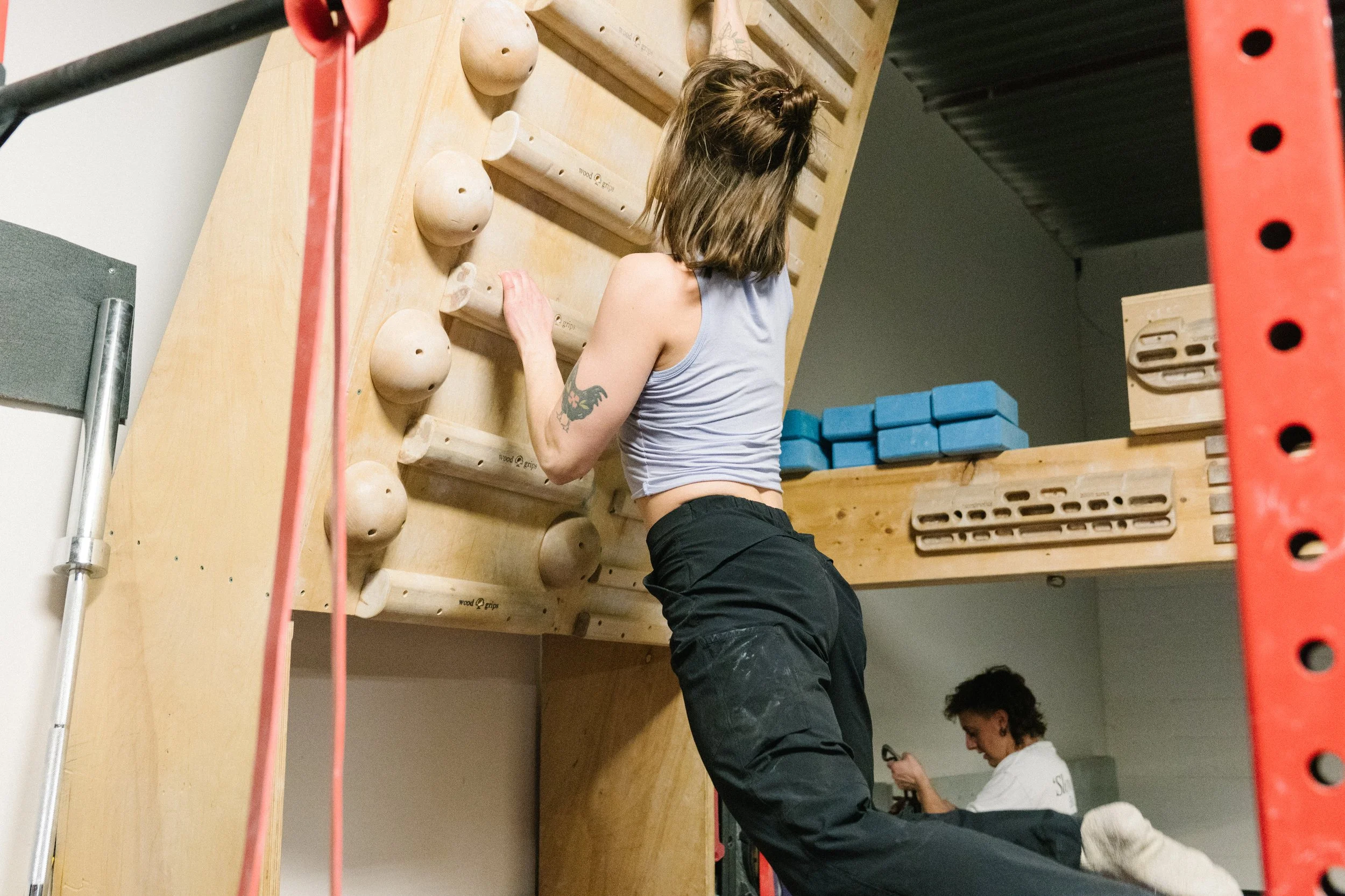 A woman with shoulder-length hair and a tattoo on her left arm is climbing an indoor rock wall. She is wearing a gray tank top and black pants. In the background, a person with curly hair and a white shirt, possibly a trainer or instructor, is looking at a device or phone.