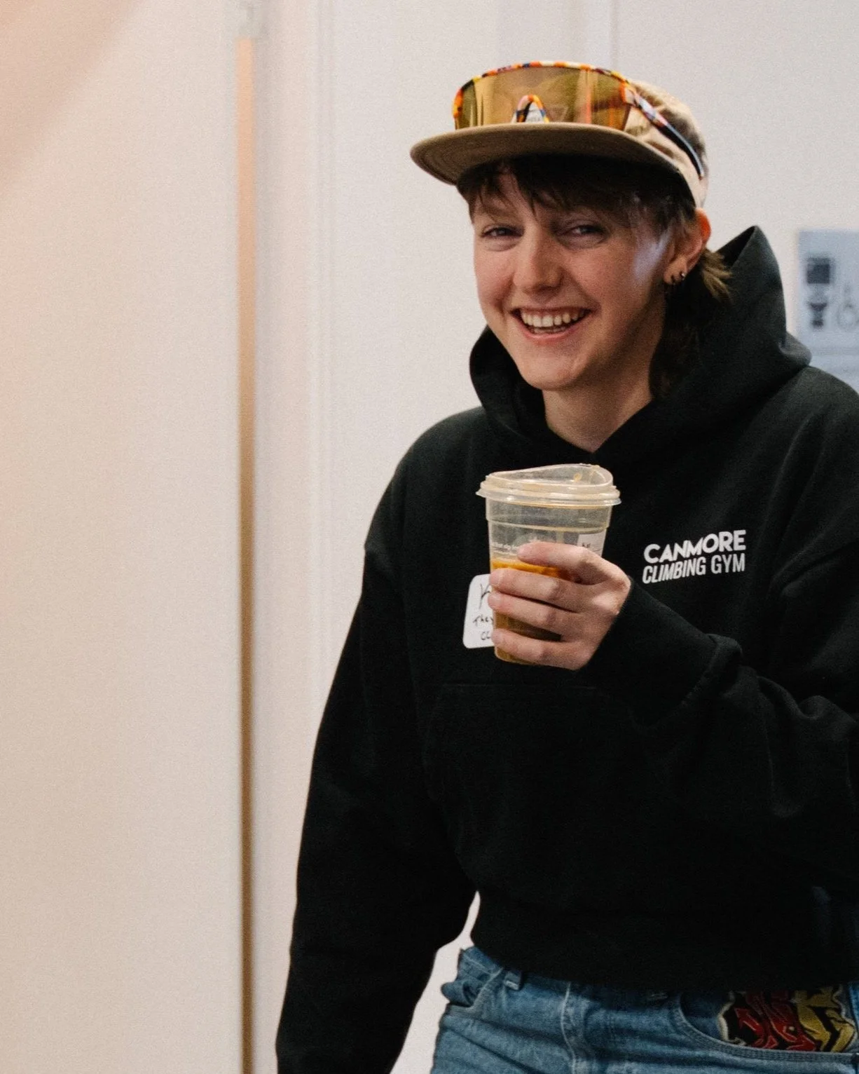 A young woman smiling, wearing a cap with sunglasses on top, a black hoodie with 'CANMORE CLIMBING GYM' written on it, and holding a plastic cup with a beverage, possibly coffee or tea.