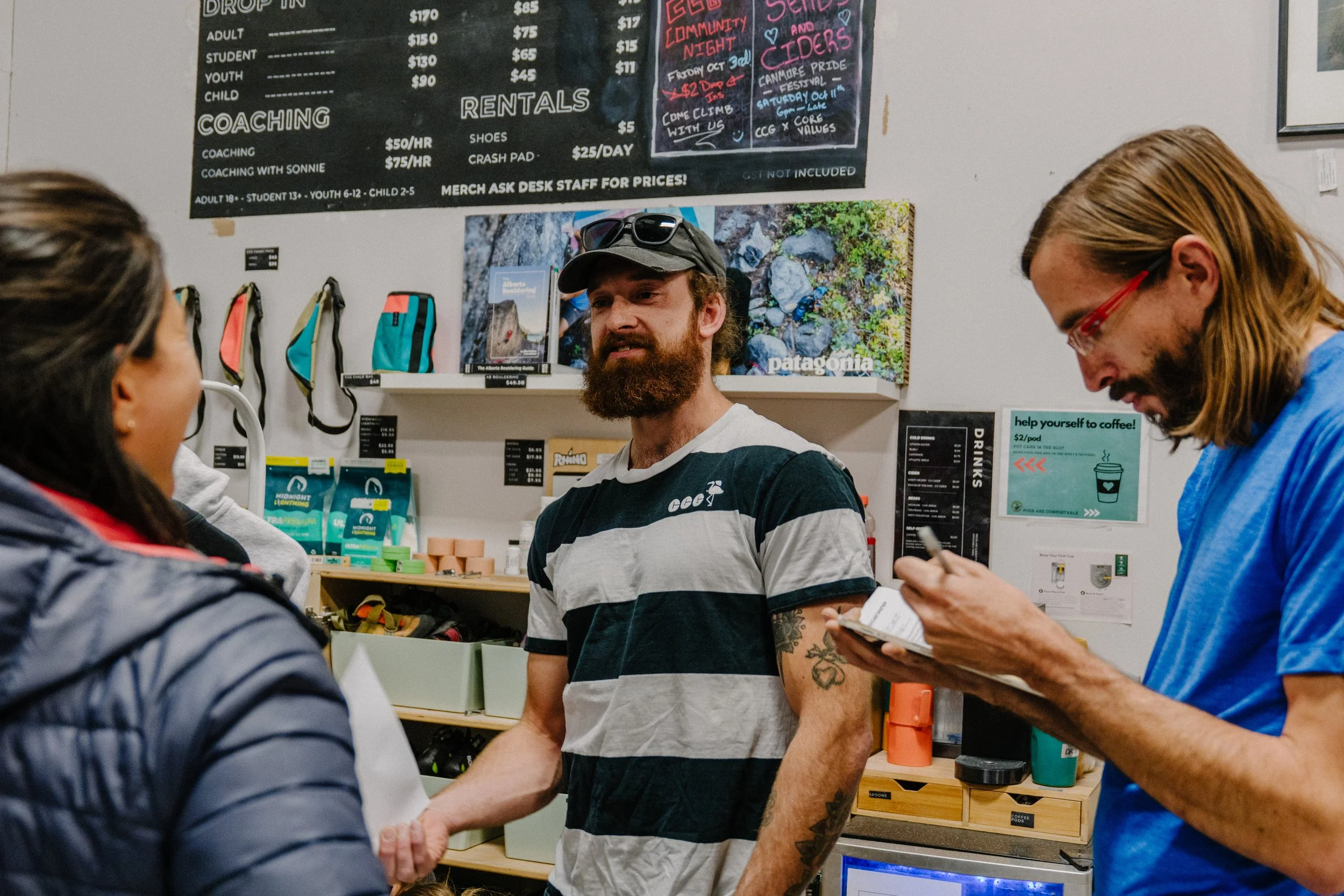 A group of people talking at a counter inside a store. There are signs and shelves with various products behind them, including a blackboard with prices and a poster for an event.