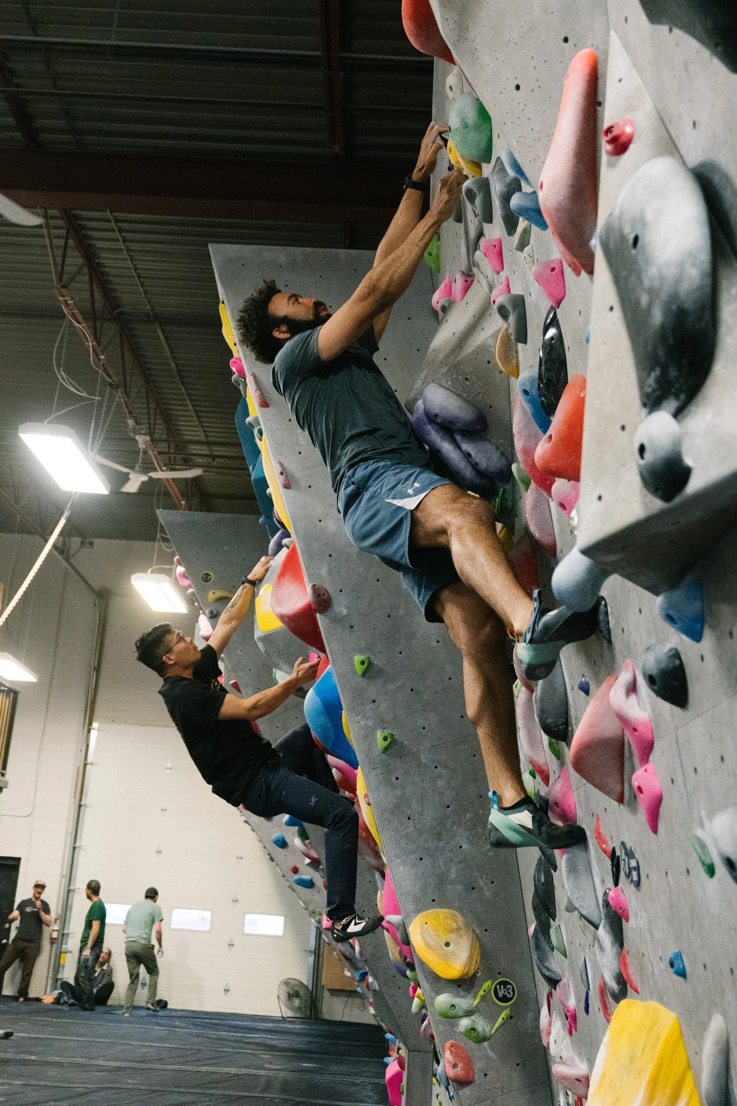 People climbing indoor bouldering wall with colorful holds, gym high ceiling, and several onlookers.