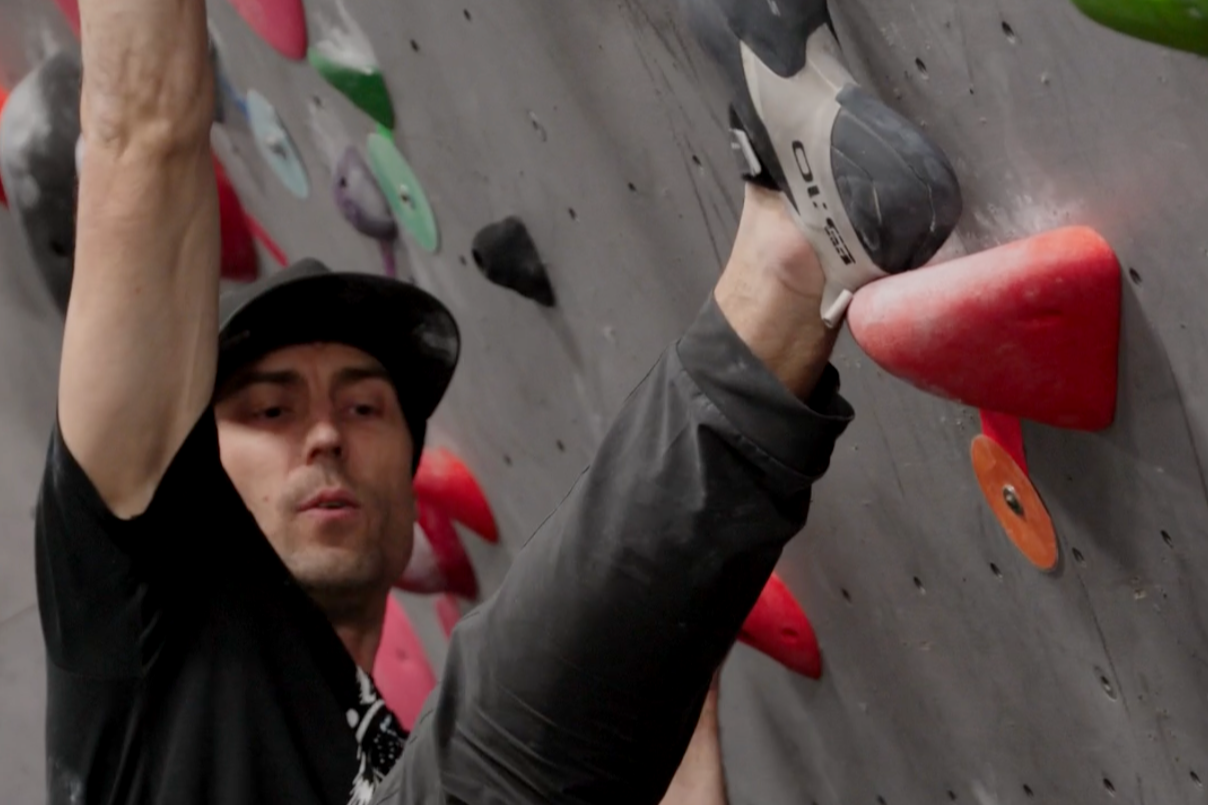 A man climbing an indoor rock wall with colorful holds, wearing a black cap and black athletic clothing.