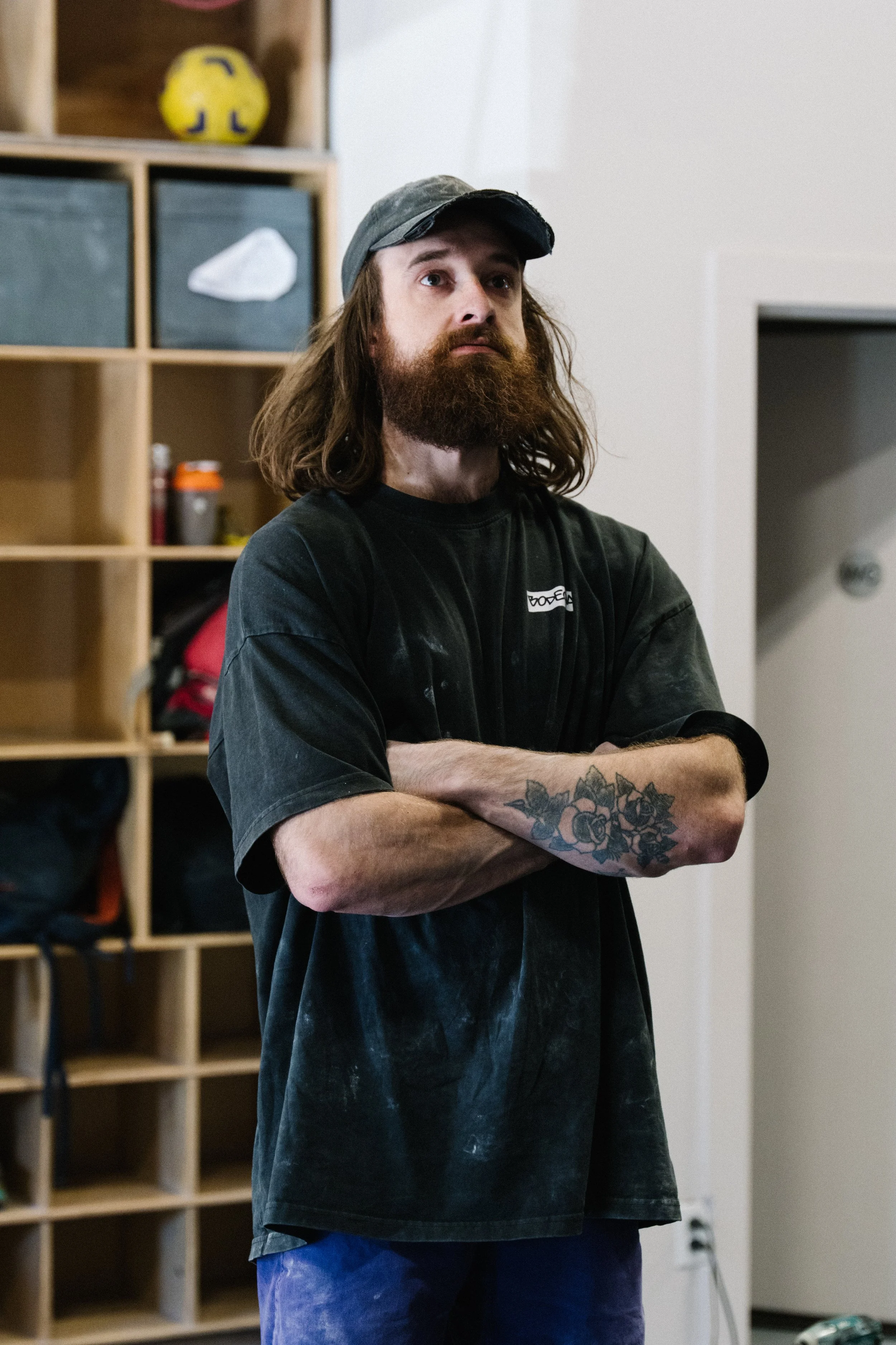 A man with long hair and a beard standing with arms crossed in a room with wooden cubby shelves in the background.