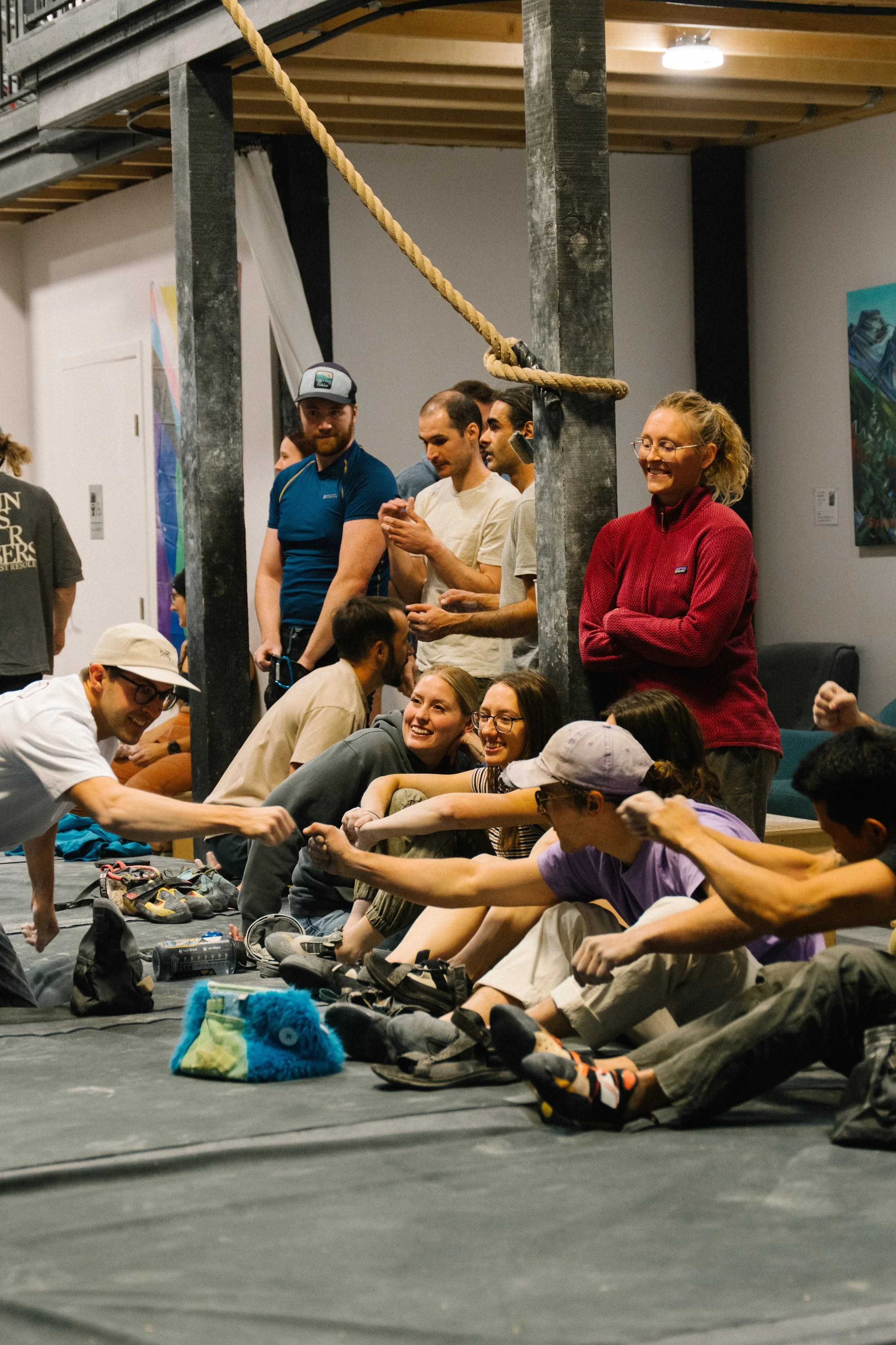 Group of people sitting and lying on the floor in an indoor space, engaging in a tug-of-war game using their hands, with some shoes and belongings placed nearby.