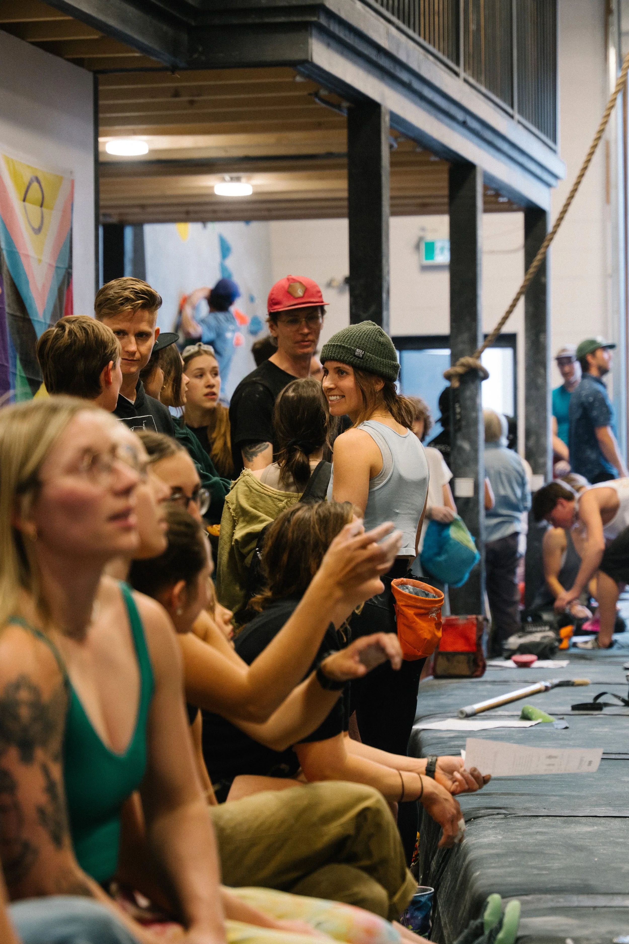 People seated on the floor participating in a yoga class or meditation session in a studio with a wooden ceiling and climbing wall in the background.