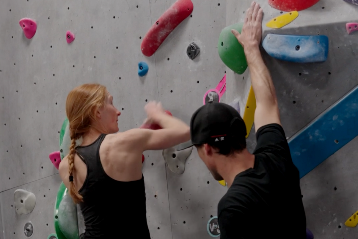 A woman with a braid is rock climbing on an indoor climbing wall, assisted by a man in a cap who is spotting her.