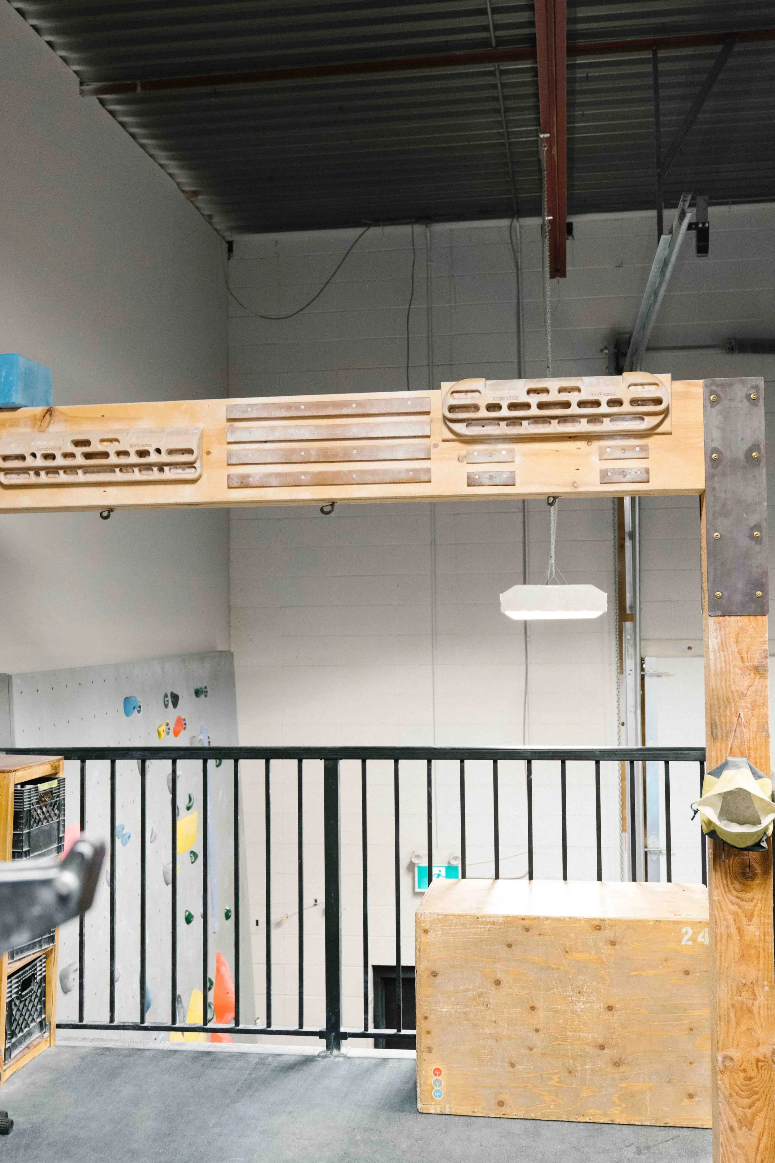 Indoor rock climbing gym with a bouldering wall decorated with climbing holds, a padded training area on top of a wooden structure, and a black metal safety railing in the foreground.
