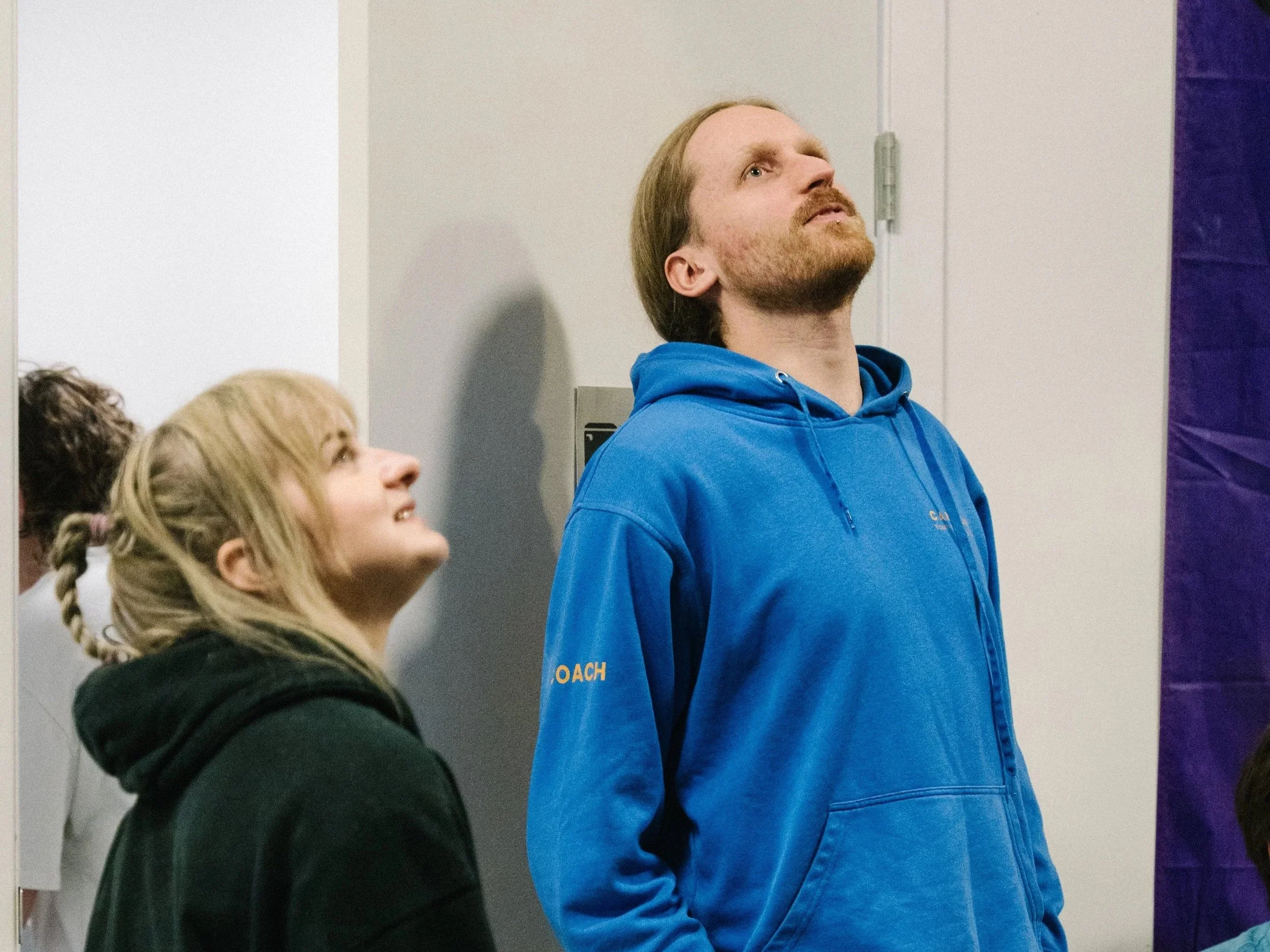 A man with a beard and long hair wearing a blue hoodie standing against a white wall, looking upward. Partially visible to the left are people with light-colored hair.