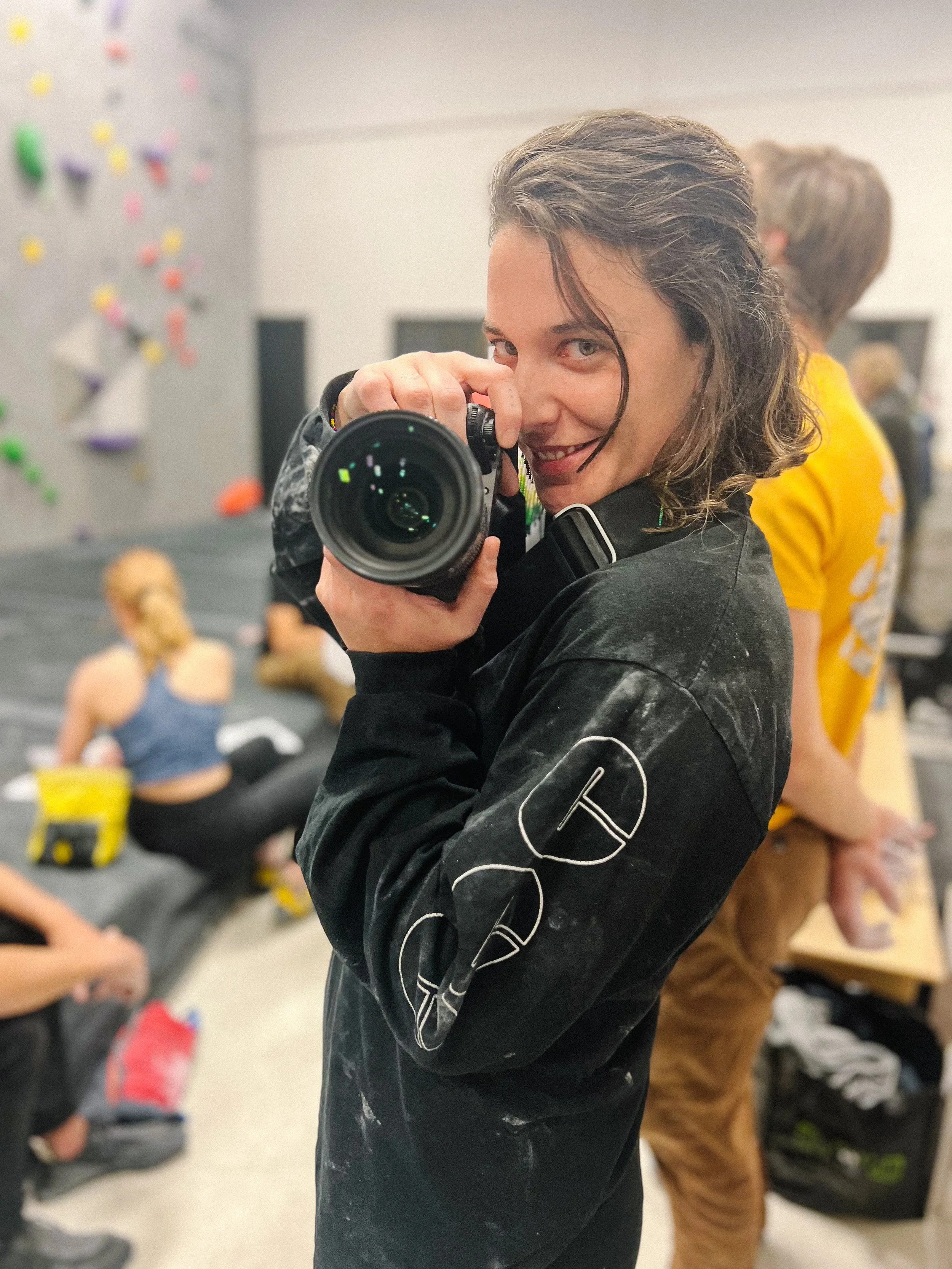 Person taking a selfie with a camera in an indoor climbing gym, with climbing holds visible on the wall in the background.