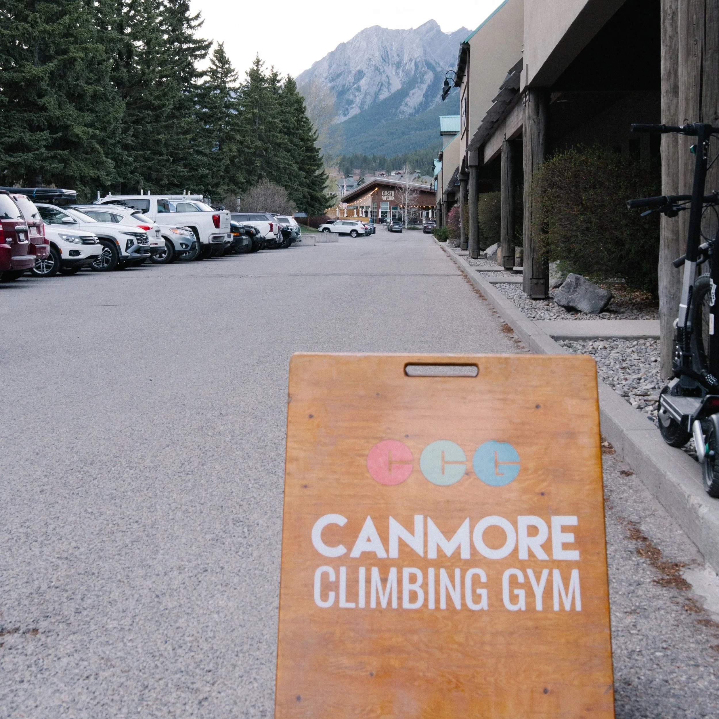 Parking lot with cars parked and a building in the background that has a sign reading "Crazy Weed". Mountain range and trees are visible in the distance.